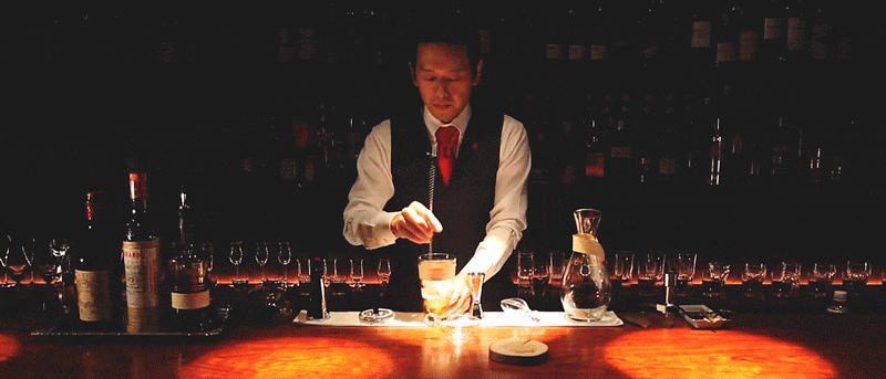 A bartender prepares a drink at a bar counter in a dimly lit setting, wearing a white shirt, black vest, and red tie, with bottles and glasses in the background.