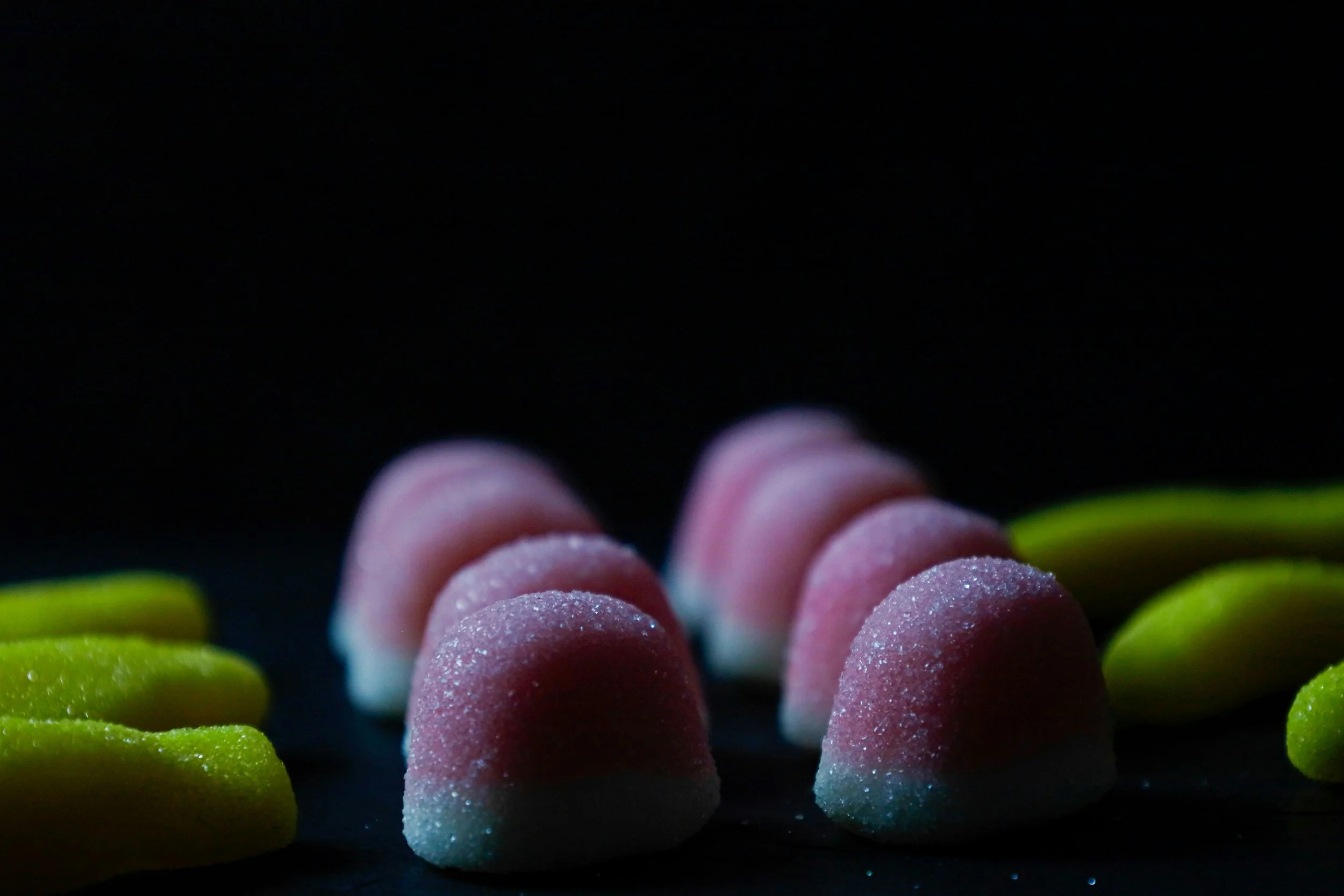 A close-up of pink and white sugar-coated gummy candies surrounded by green candies on a dark background.