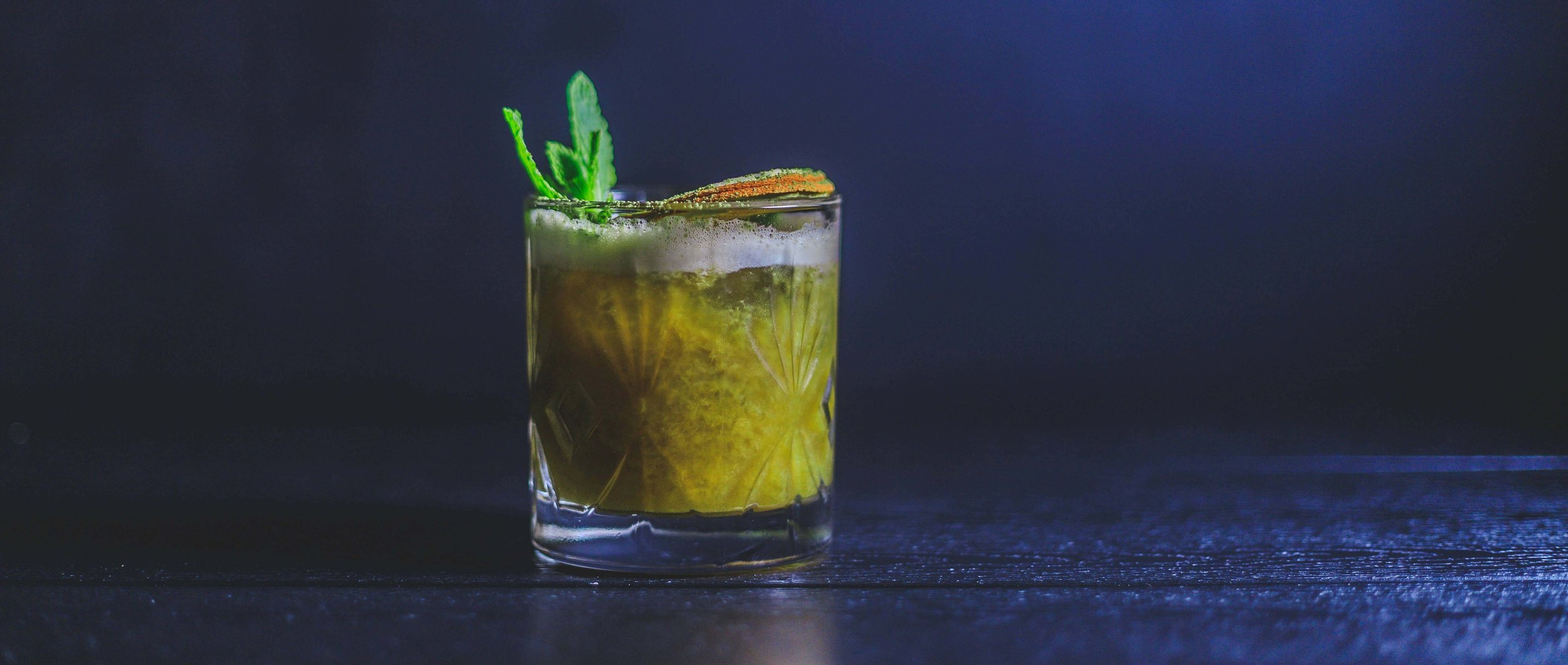 A glass of cocktail with green garnish and a slice of lime, set against a dark background.