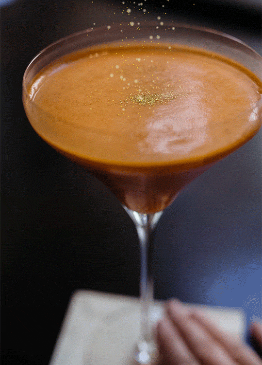A cocktail in a martini glass with a smooth, frothy orange-brown surface, garnished with gold glitter, against a dark background.