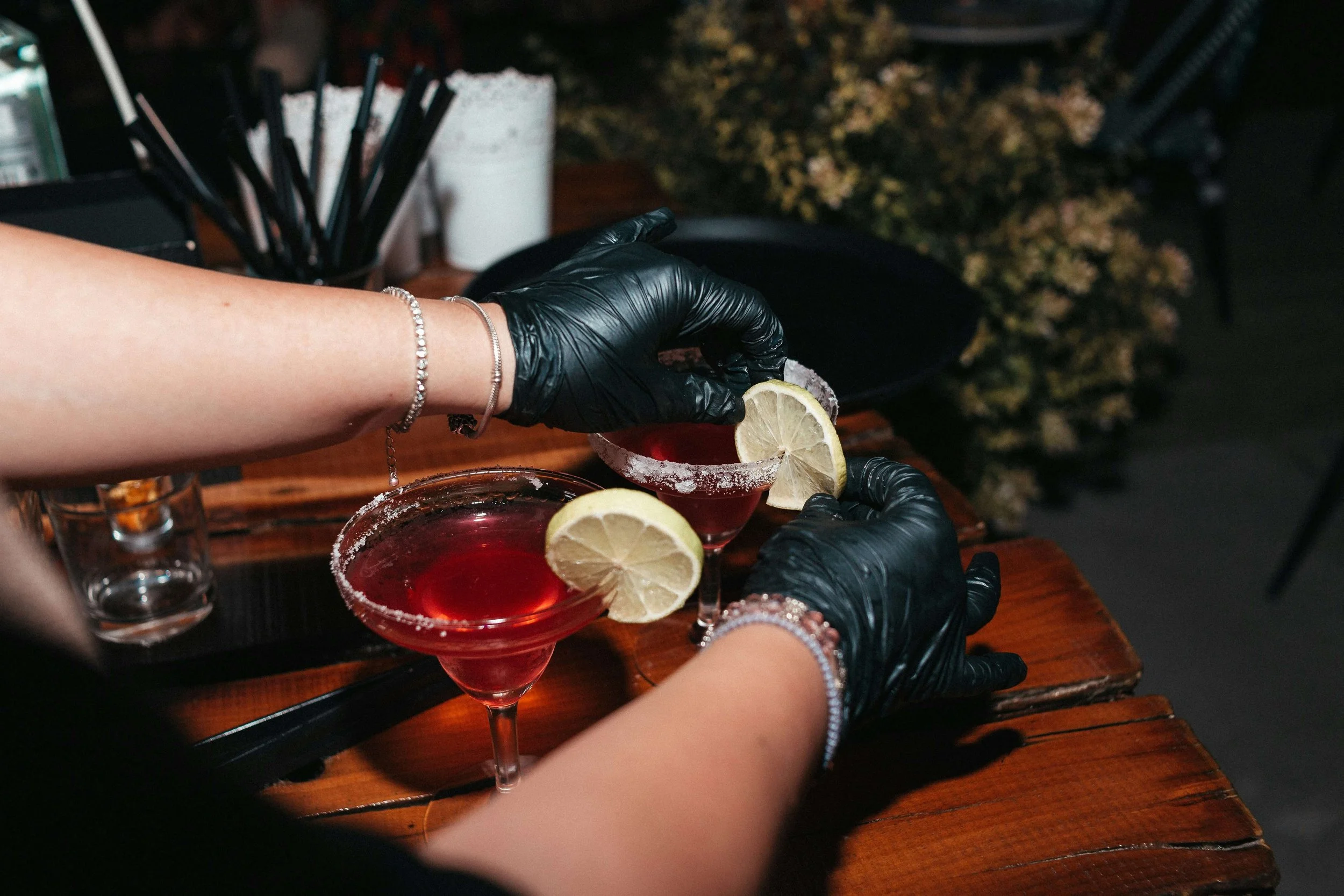 Two people wearing black gloves are garnishing red cocktails with lemon slices at a bar.