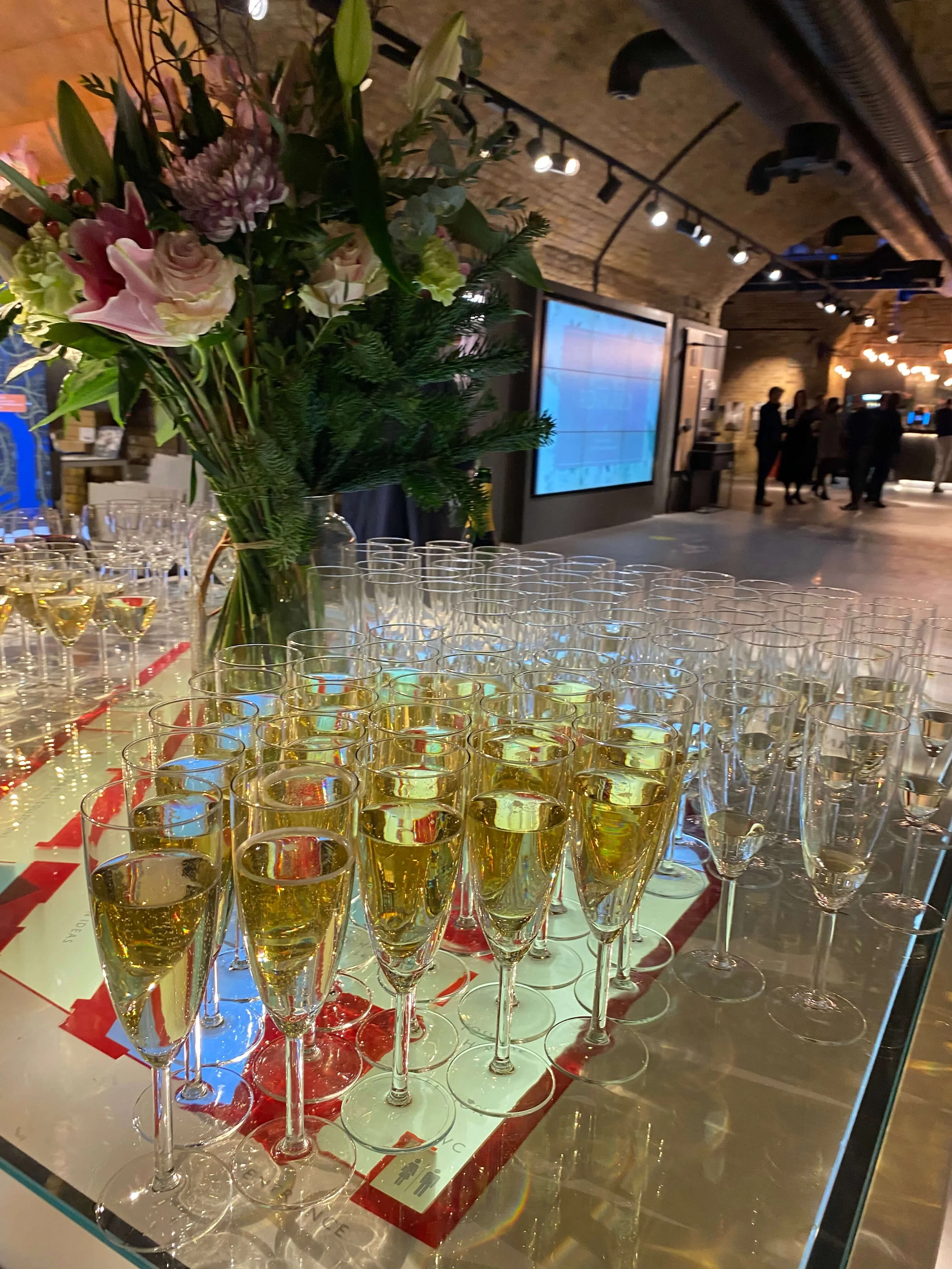 A display of champagne glasses filled with a bubbly drink and a tall bouquet of pink and purple flowers on a table at an event venue with people in the background.