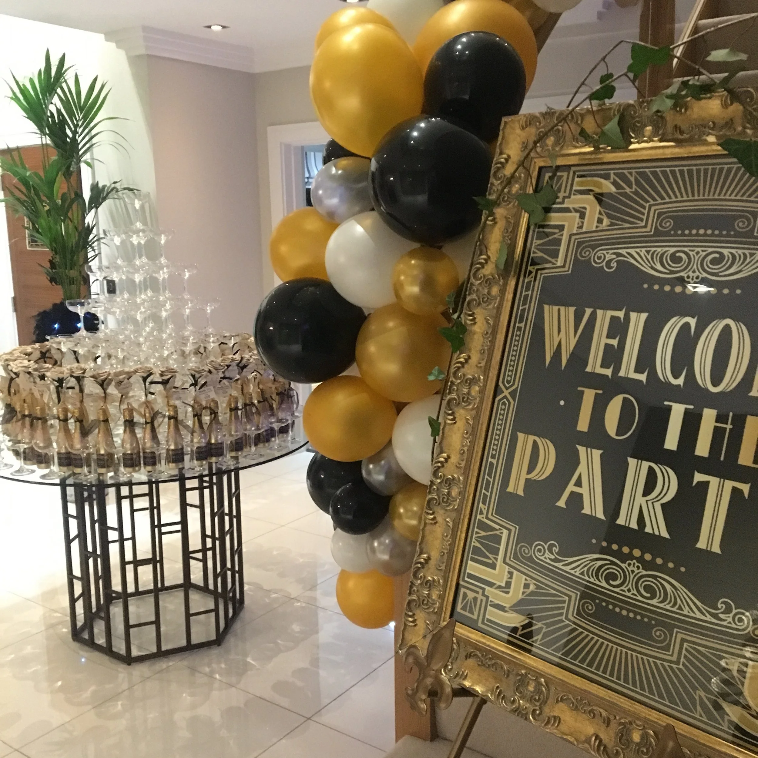 Decorative party setup with a black, gold, white, and gray balloon arch, a champagne glass pyramid, and a 'Welcome to the Party' sign in an ornate gold frame.