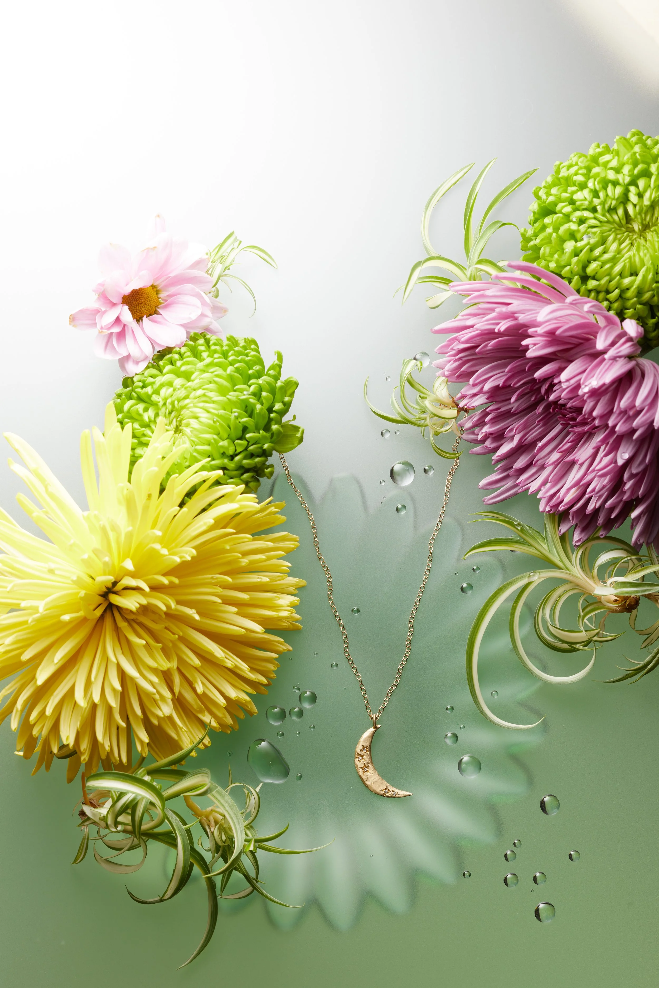 A silver crescent moon necklace with star accents hanging among colorful flowers including pink, yellow, and purple chrysanthemums and green spider plants, with water droplets on a reflective surface.
