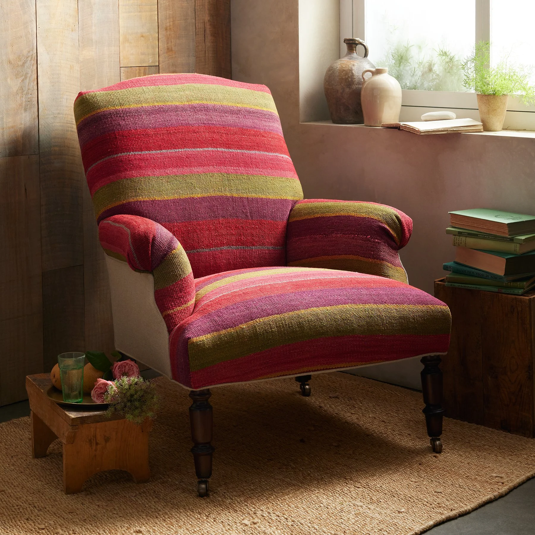 A colorful striped armchair with wooden legs and wheels in a cozy room near a window, with a small wooden side table holding a glass of green liquid, pink roses, and greenery, and a stack of books nearby.