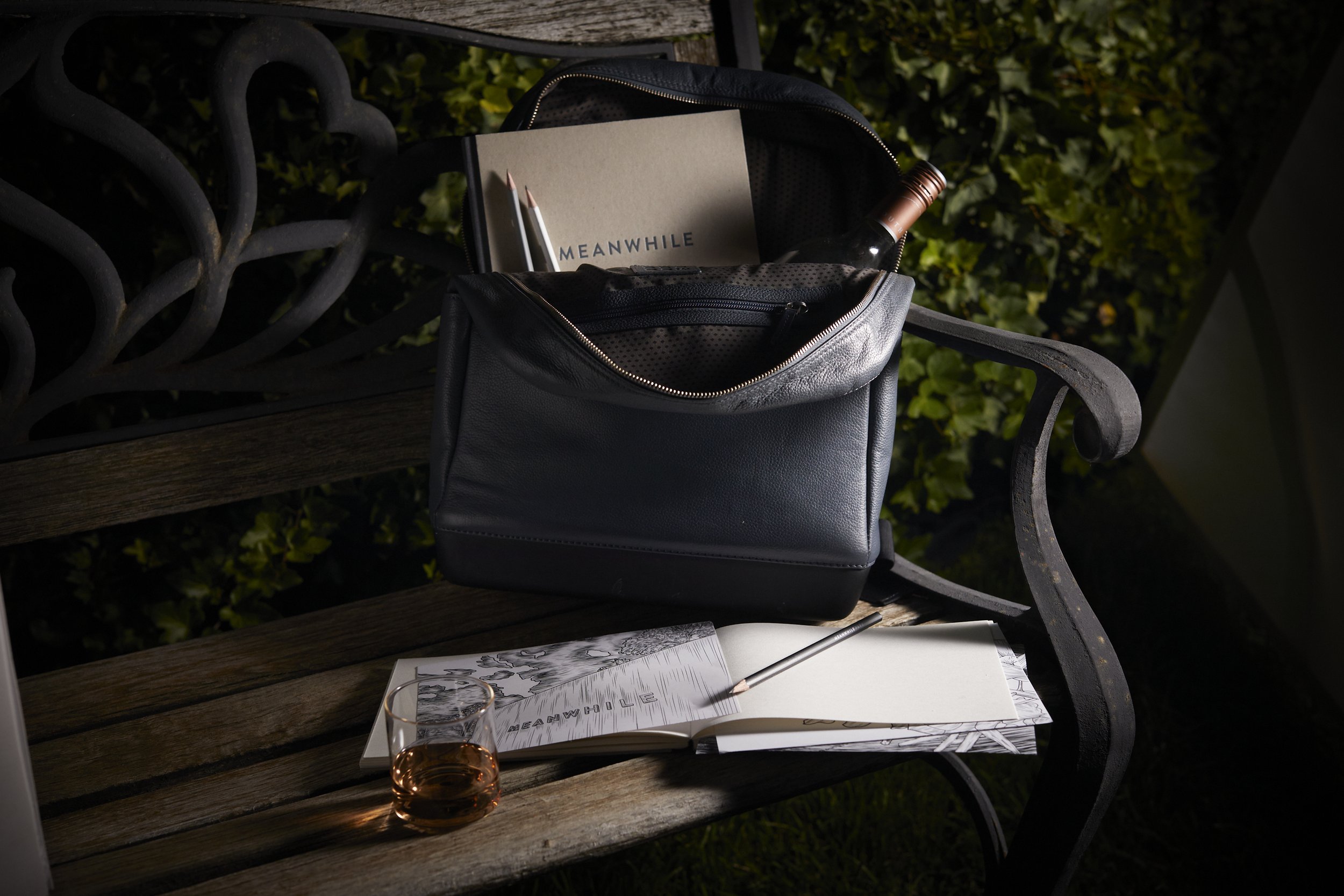 A black leather bag on a wooden bench contains a book, two pencils, and a wine bottle. On the bench, there's an open sketchbook with a pencil on it and a glass of rosé wine.