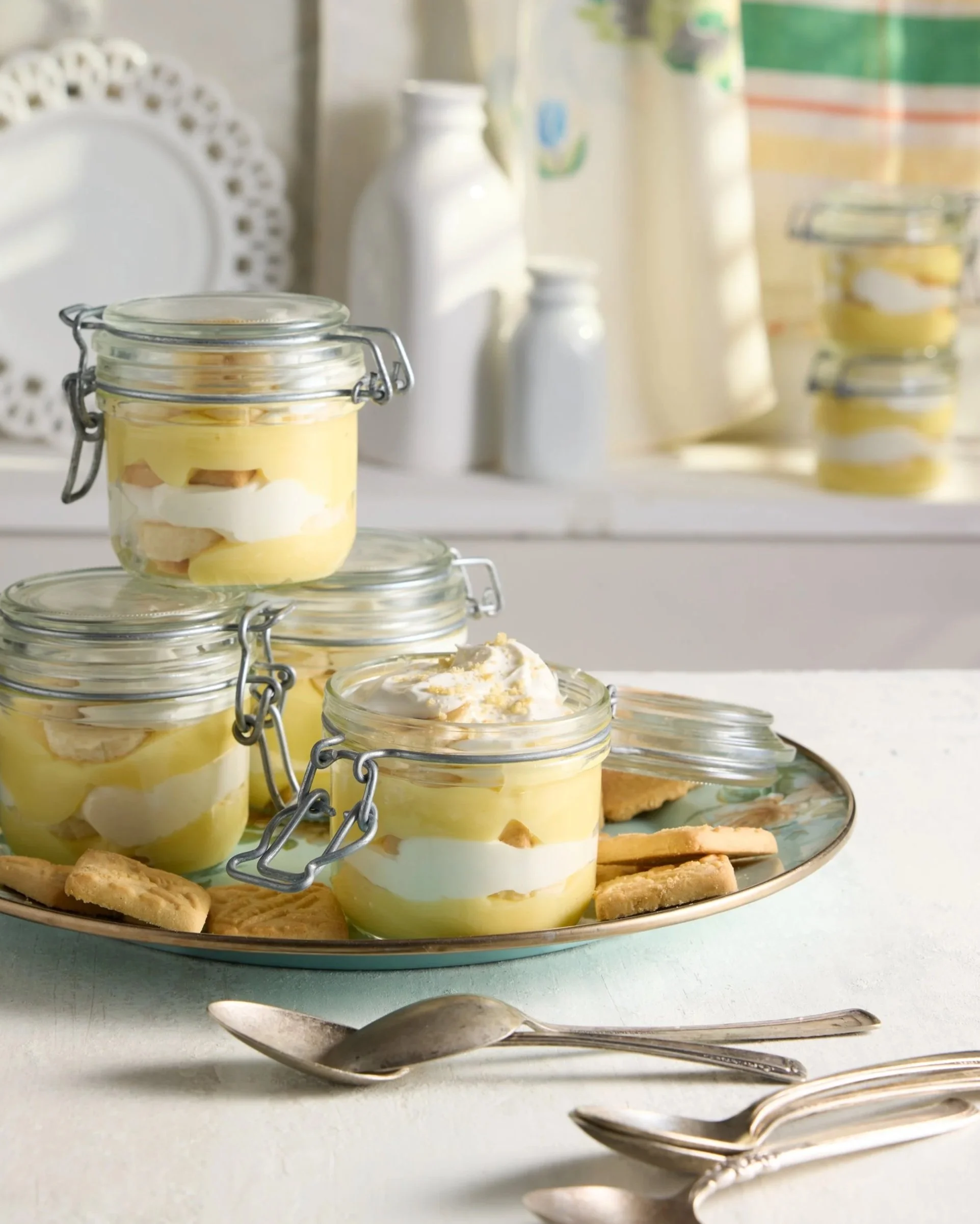 Individual lemon trifles in small glass jars with cookies on a decorative plate, set on a white table with silver spoons.