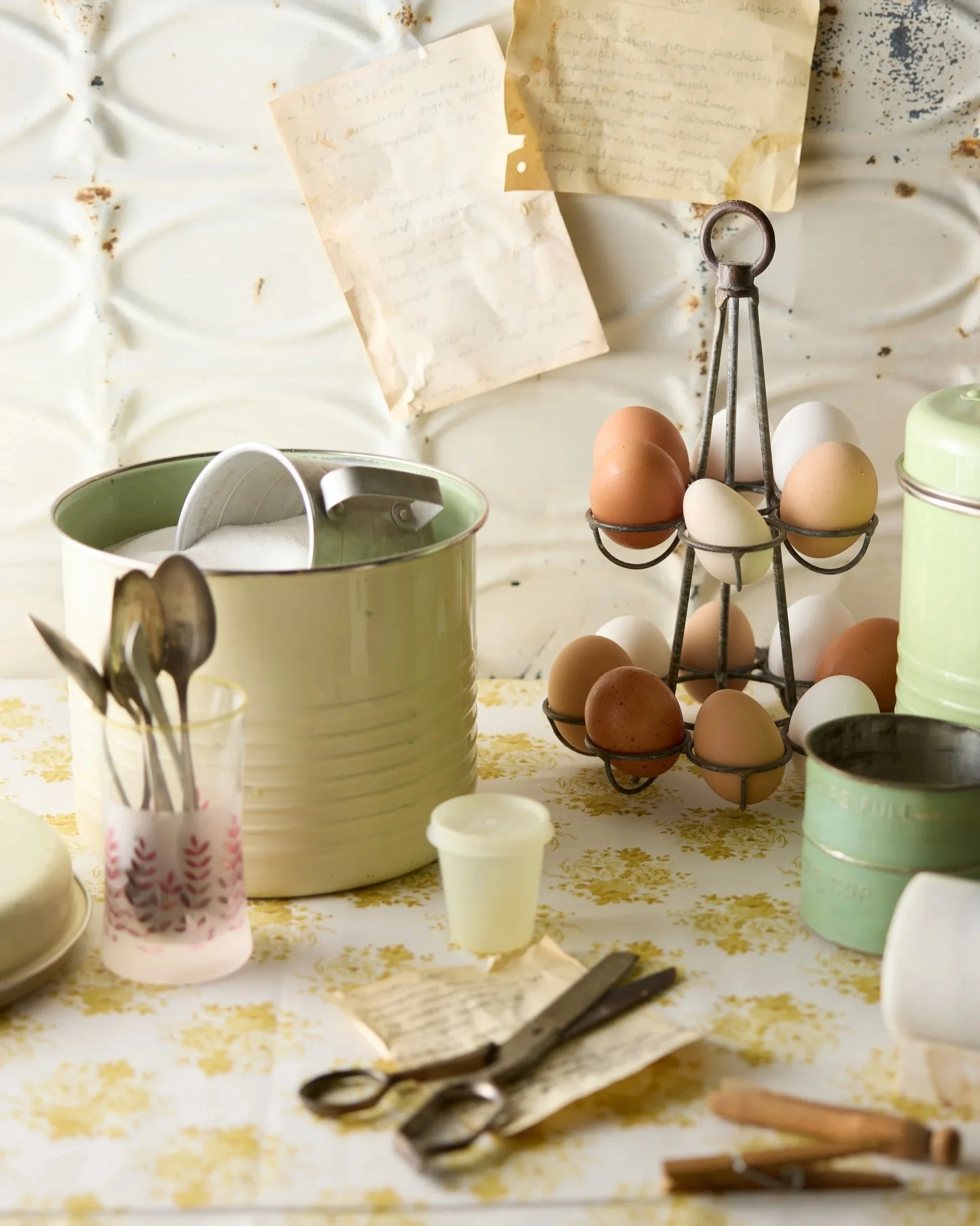 A vintage-style kitchen or pantry scene with a table covered in a floral cloth, holding eggs in a metal egg holder, metal utensils in a pink floral cup, scissors, a small jar, and metal tins, with handwritten notes or recipes taped on a white wall or backsplash in the background.