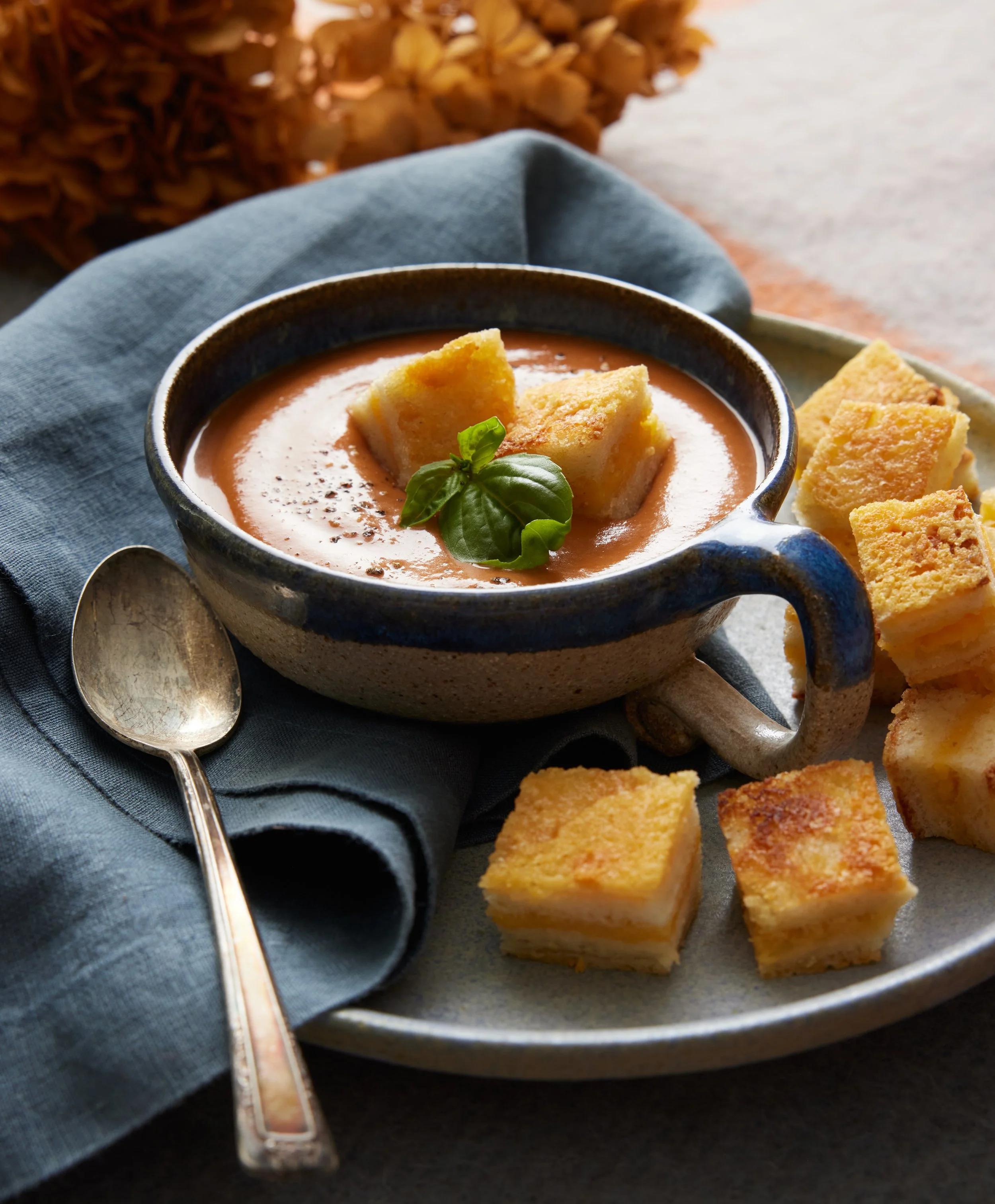 A rustic ceramic bowl of tomato soup garnished with basil and croutons, surrounded by squares of cornbread on a gray plate with a silver spoon and blue napkin.