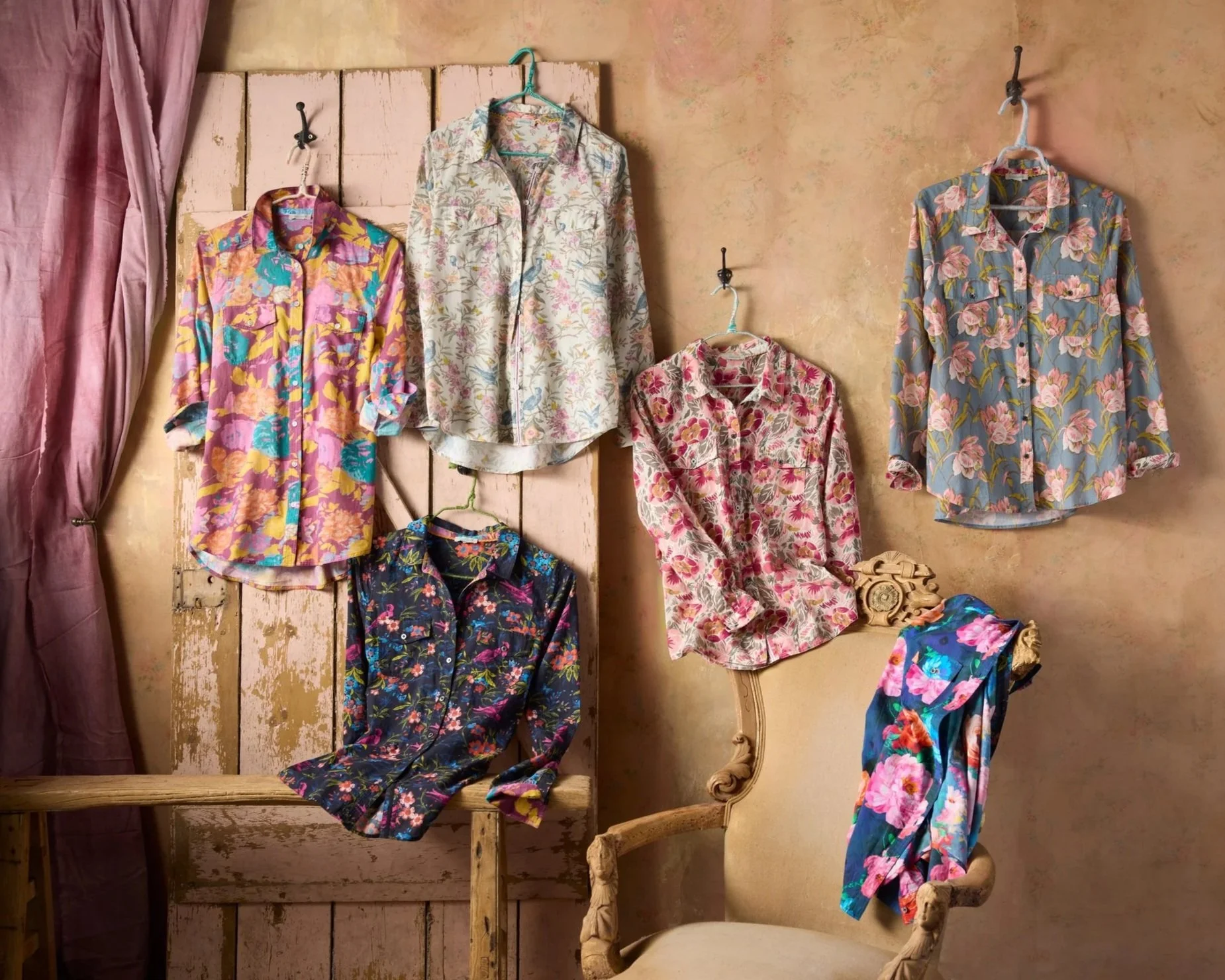 Multiple floral-patterned shirts hanging on hooks and a chair in a vintage-style room with pink and beige walls.