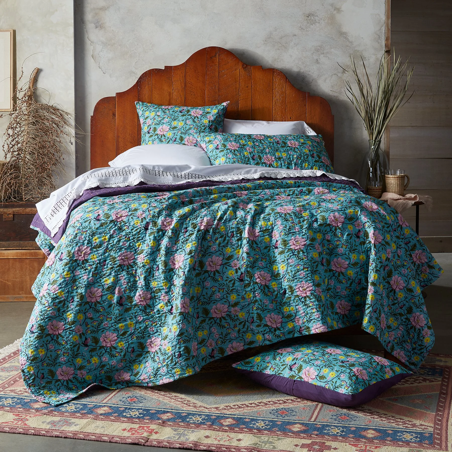 A bed with a floral quilt, matching pillowcases, and white sheets in a bedroom with a wooden headboard, a dried plant on the left, and a vase with tall grasses on the right, placed on a patterned rug.