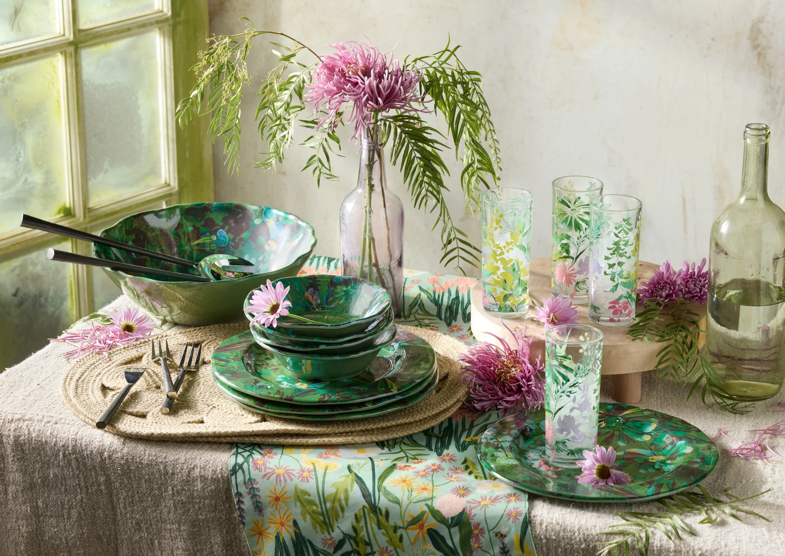 A table setting with green floral dinnerware, pink flowers, and a glass vase with pink flowers, positioned near a window with sunlight. The table has a beige textured tablecloth, and there are colorful glassware and silverware arranged on it.