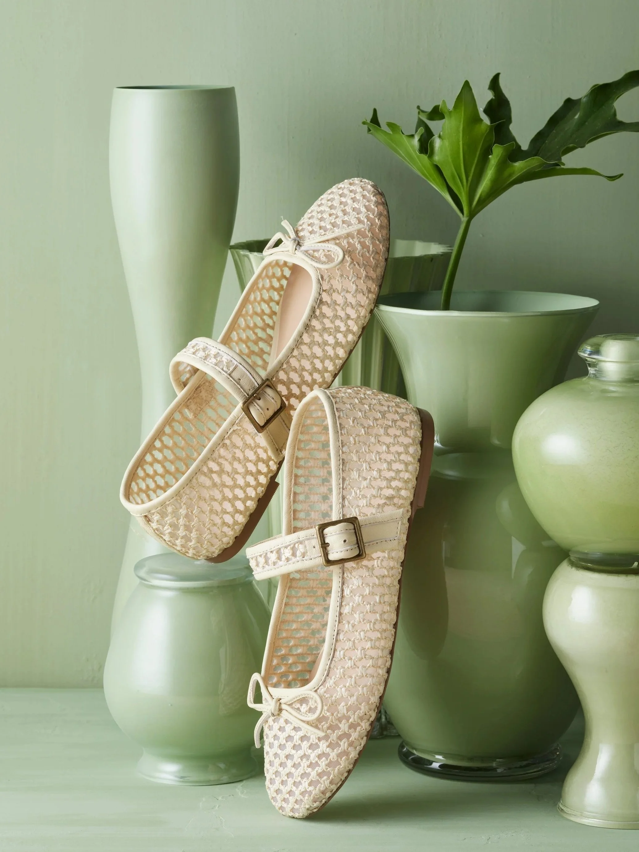 Beige woven ballet flats with ankle straps, surrounded by various green vases and a potted plant.