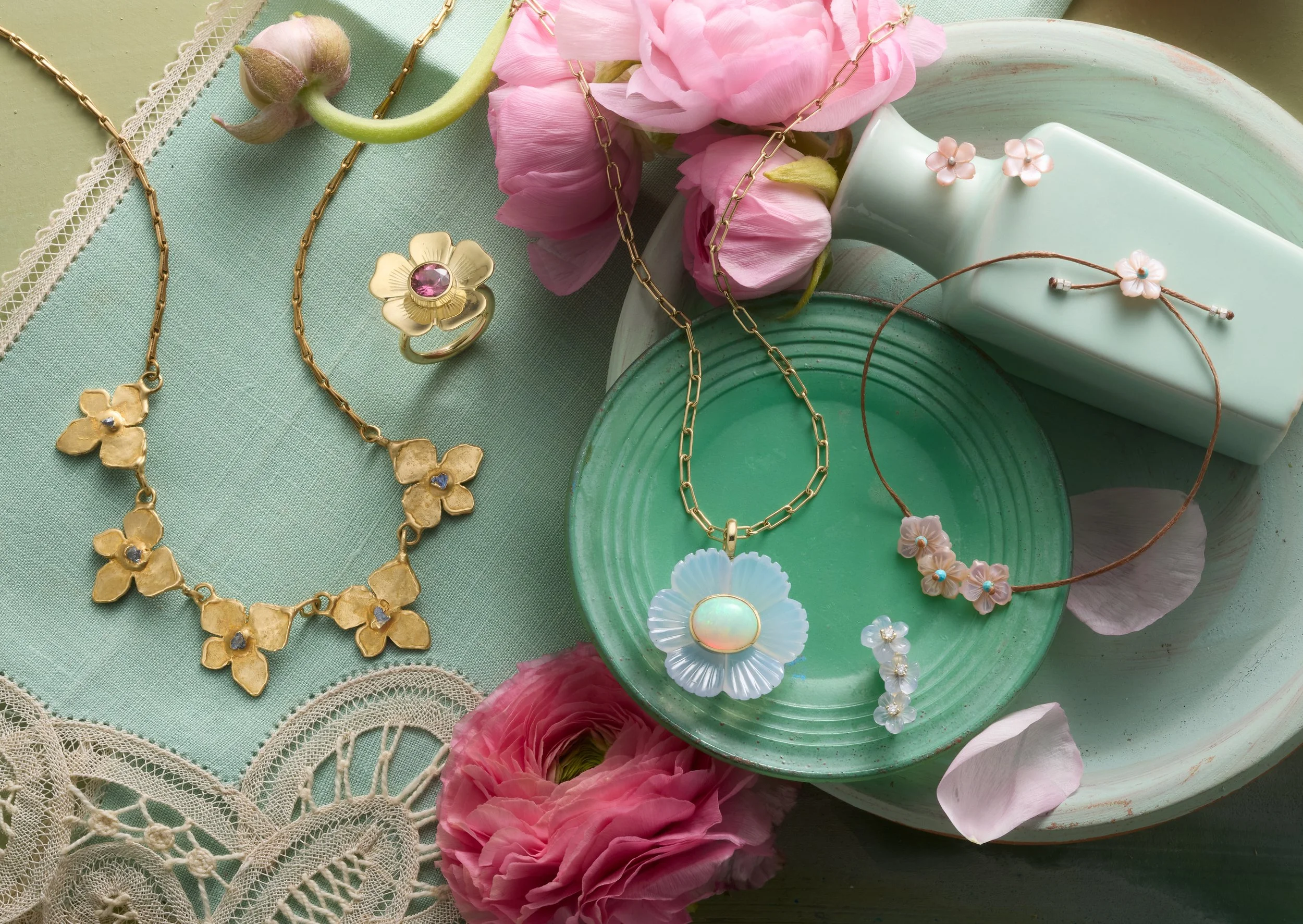 A collection of jewelry, including necklaces, rings, bracelets, and earrings, displayed on a green tray and a lace cloth, surrounded by pink flowers and a round soap or decorative box.