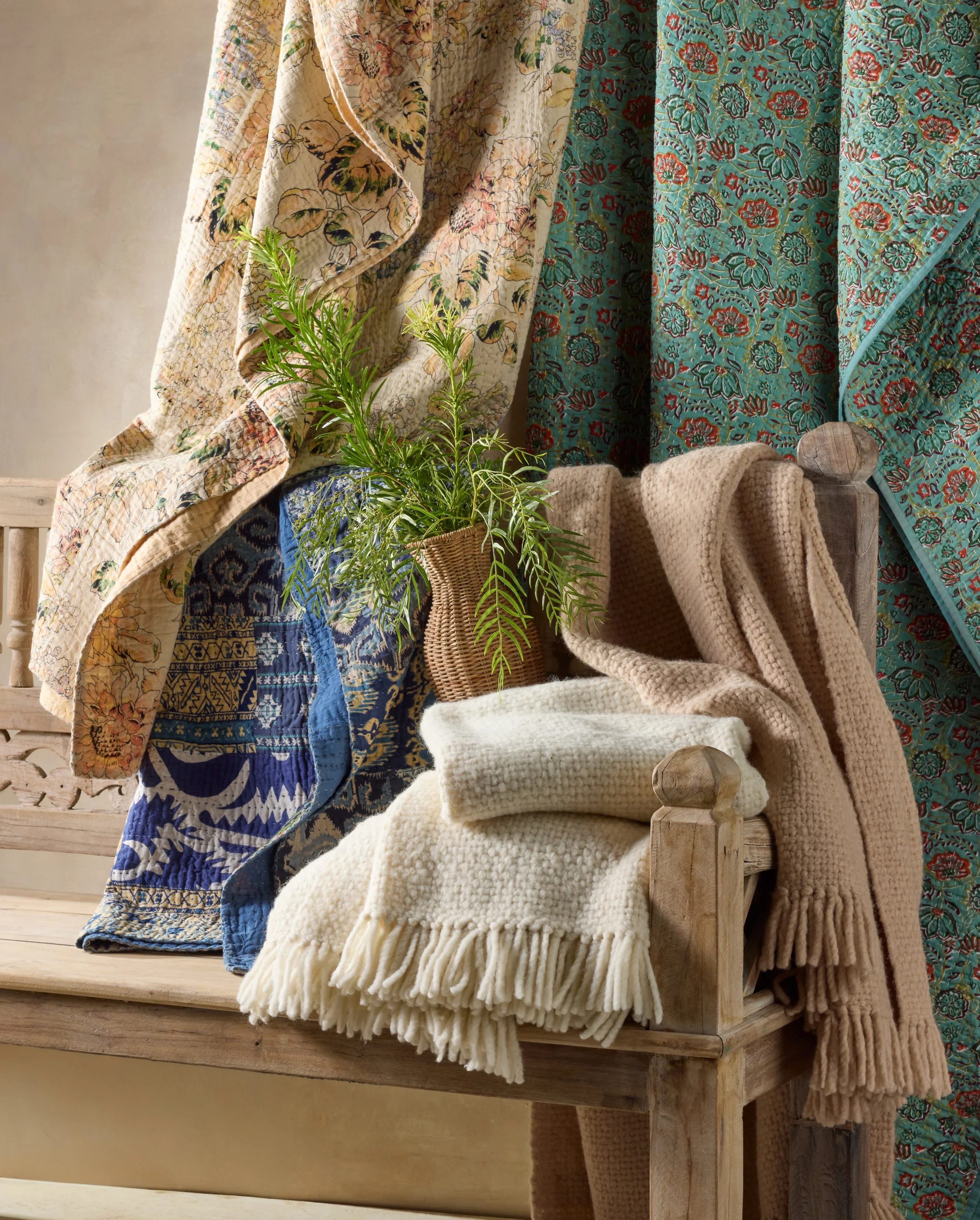 A wooden bench with beige, white, and tan blankets, a potted plant, and colorful floral fabrics hanging in the background.