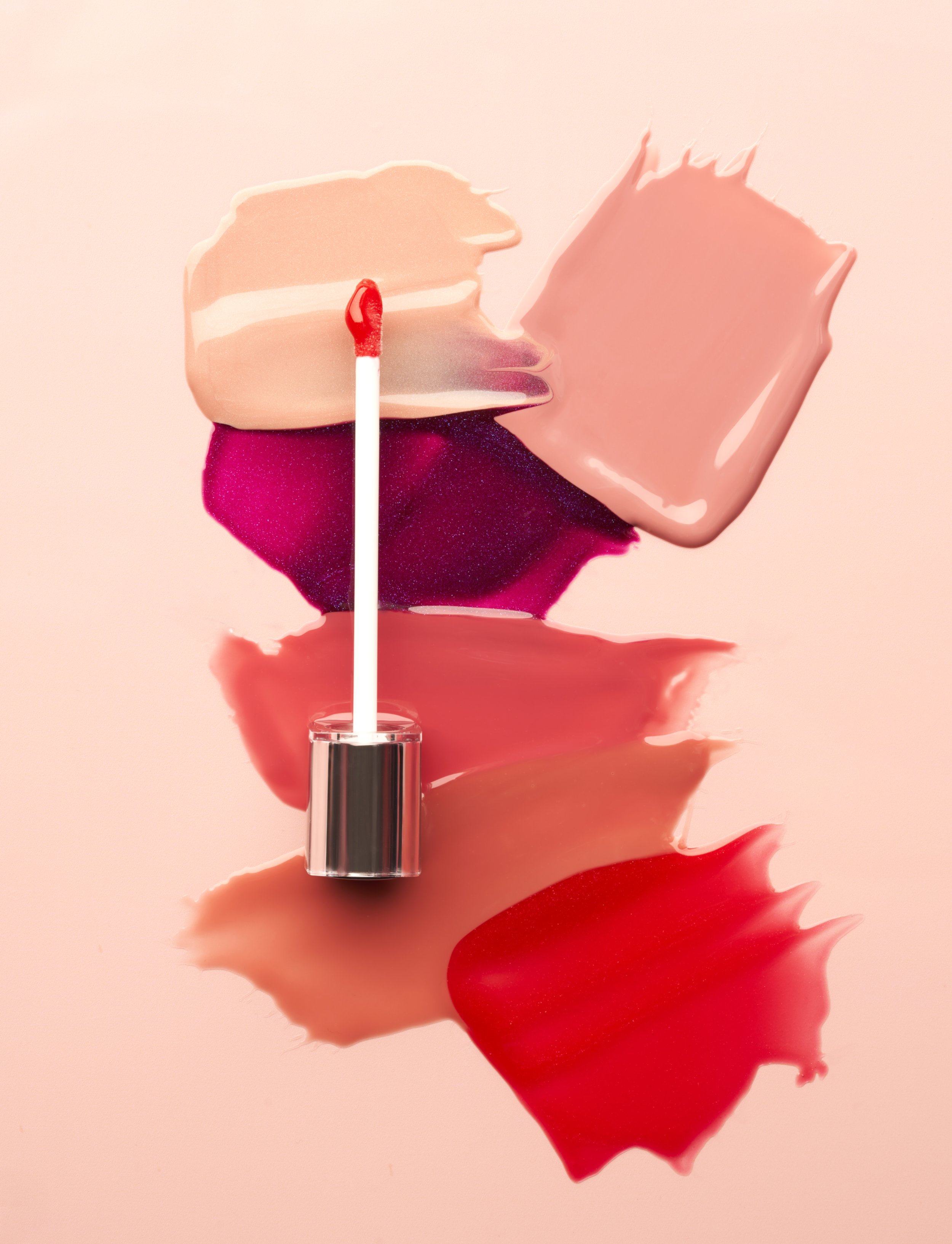 A makeup lipstick lying on a background with pink, red, and beige swirls, with a small red-tipped applicator brush standing upright in the lipstick.