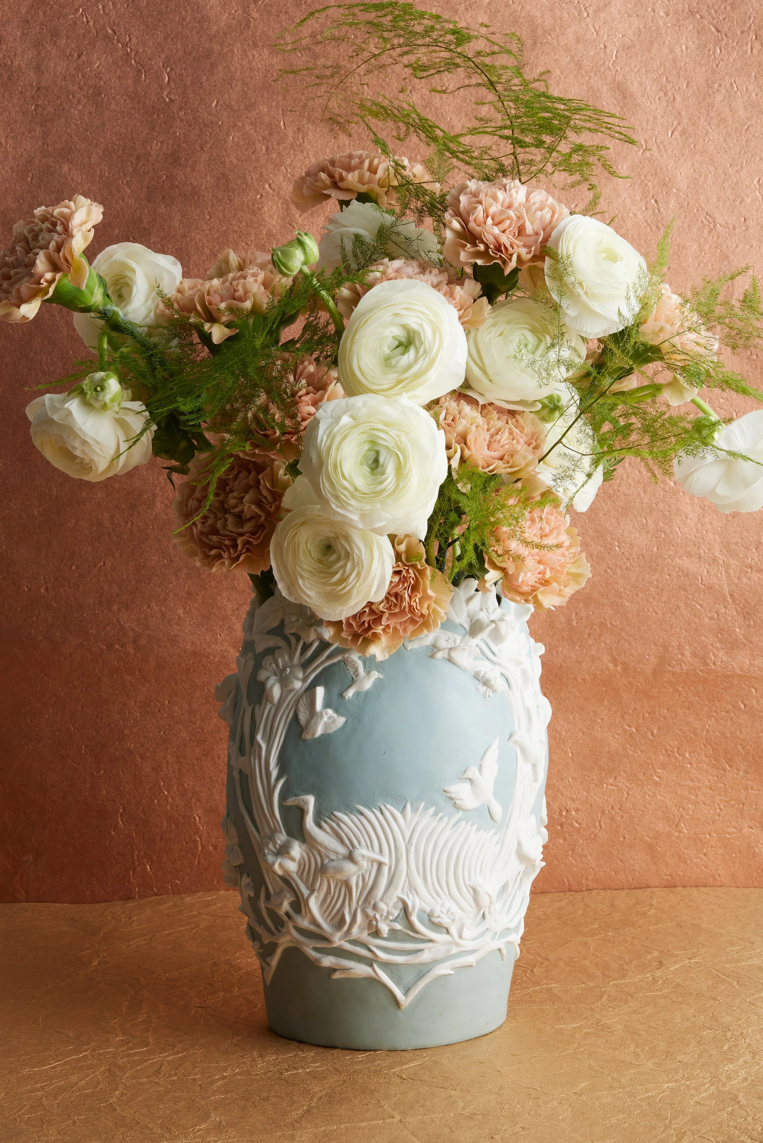 A decorative ceramic vase with intricate white leaf and swirl patterns, holding a bouquet of cream-colored and pink carnations, white ranunculus, and green fern leaves, set against a textured peach background.
