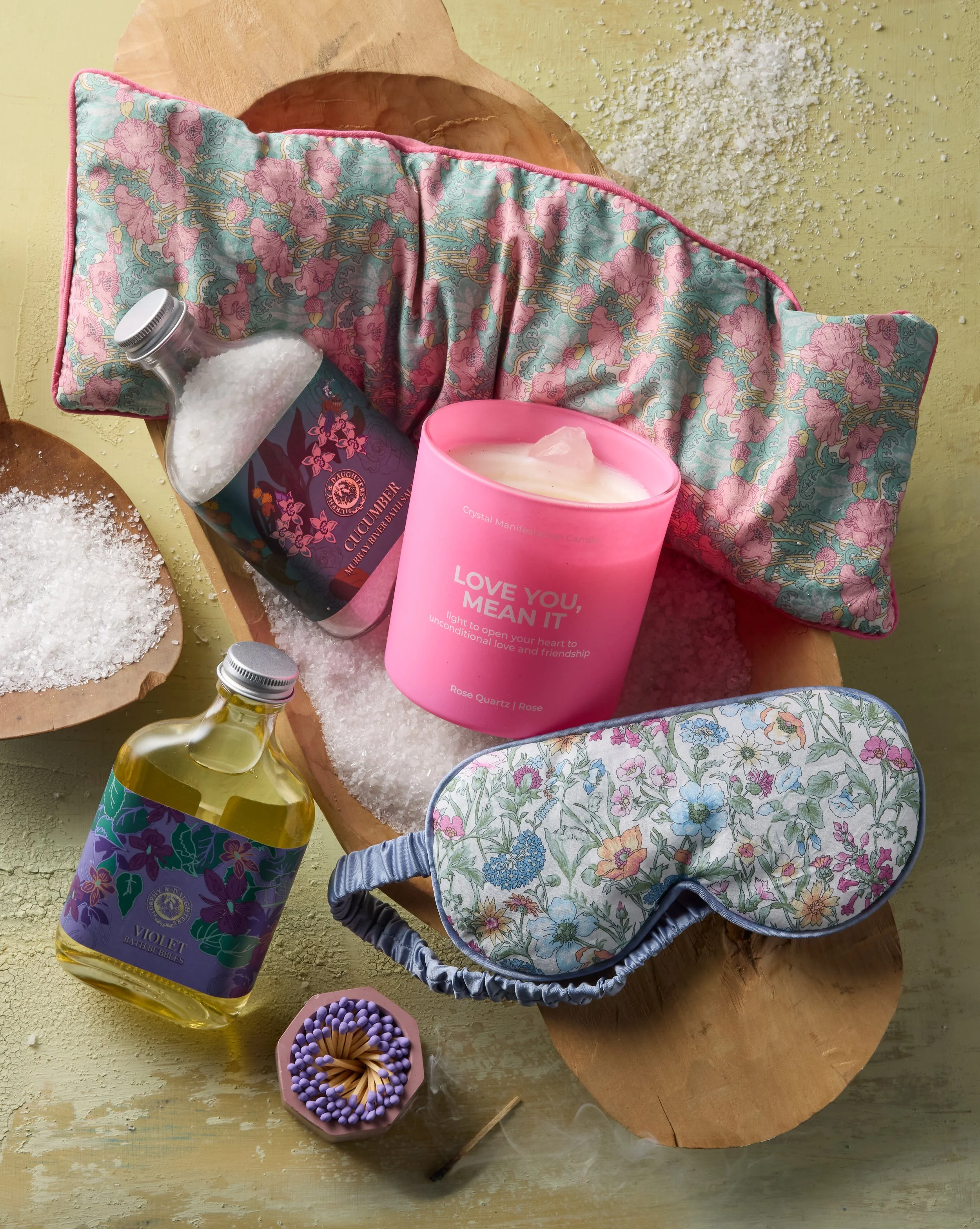 A spa or relaxation setup on a wooden surface featuring a bath pillow with pink and blue floral pattern, a pink candle with the words 'Love you, mean it,' a bottle of cucumber shower gel, a bottle of violet bath bubbles, a floral sleep mask, a container of bath salts, a matchstick, and a small dish of scattered bath salts.