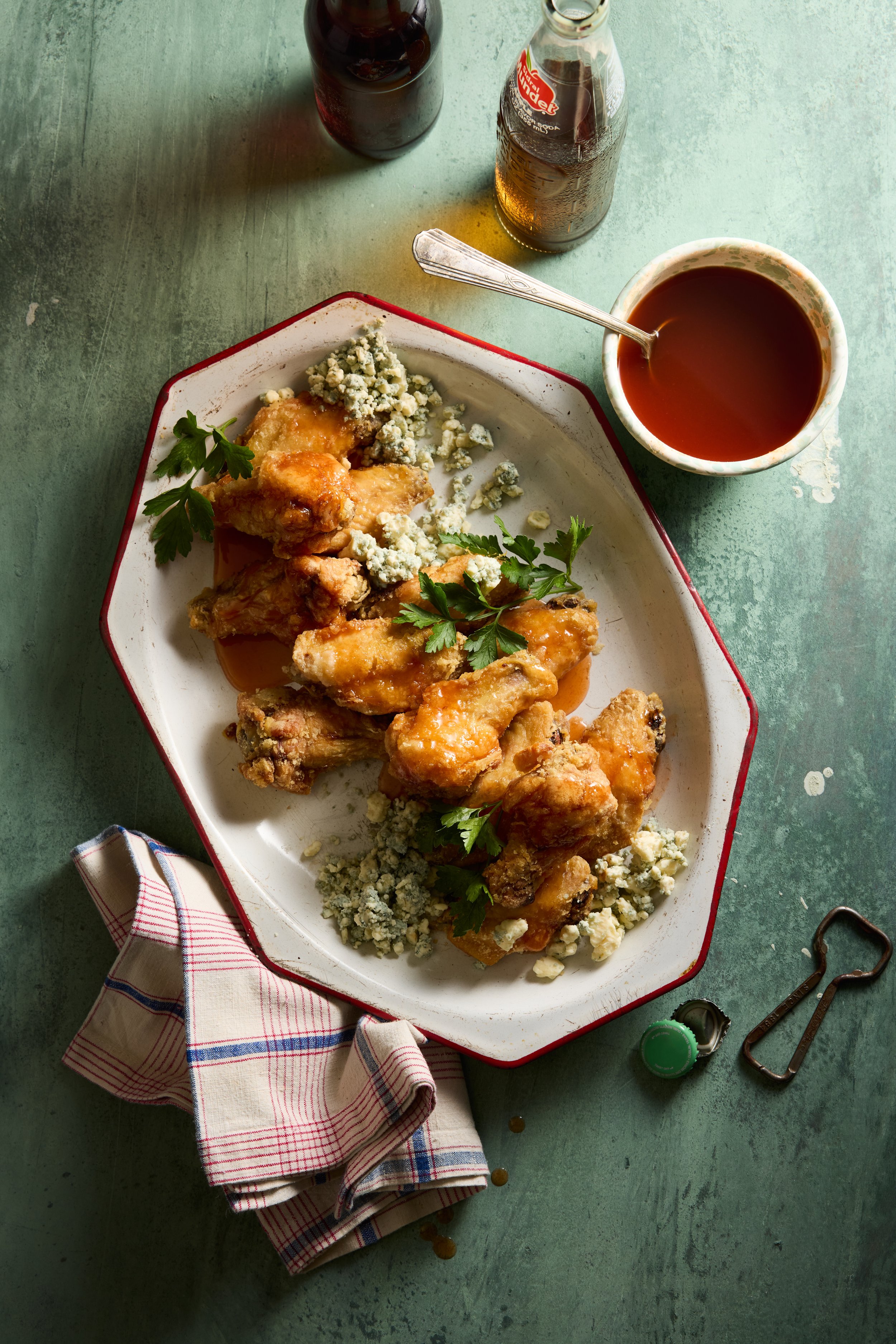 Fried chicken wings on a white platter garnished with blue cheese crumbles and parsley, accompanied by a bowl of hot sauce or dressing on a green table, with a checkered napkin, a bottle of hot sauce, an open bottle of beer, and a small bottle cap ne