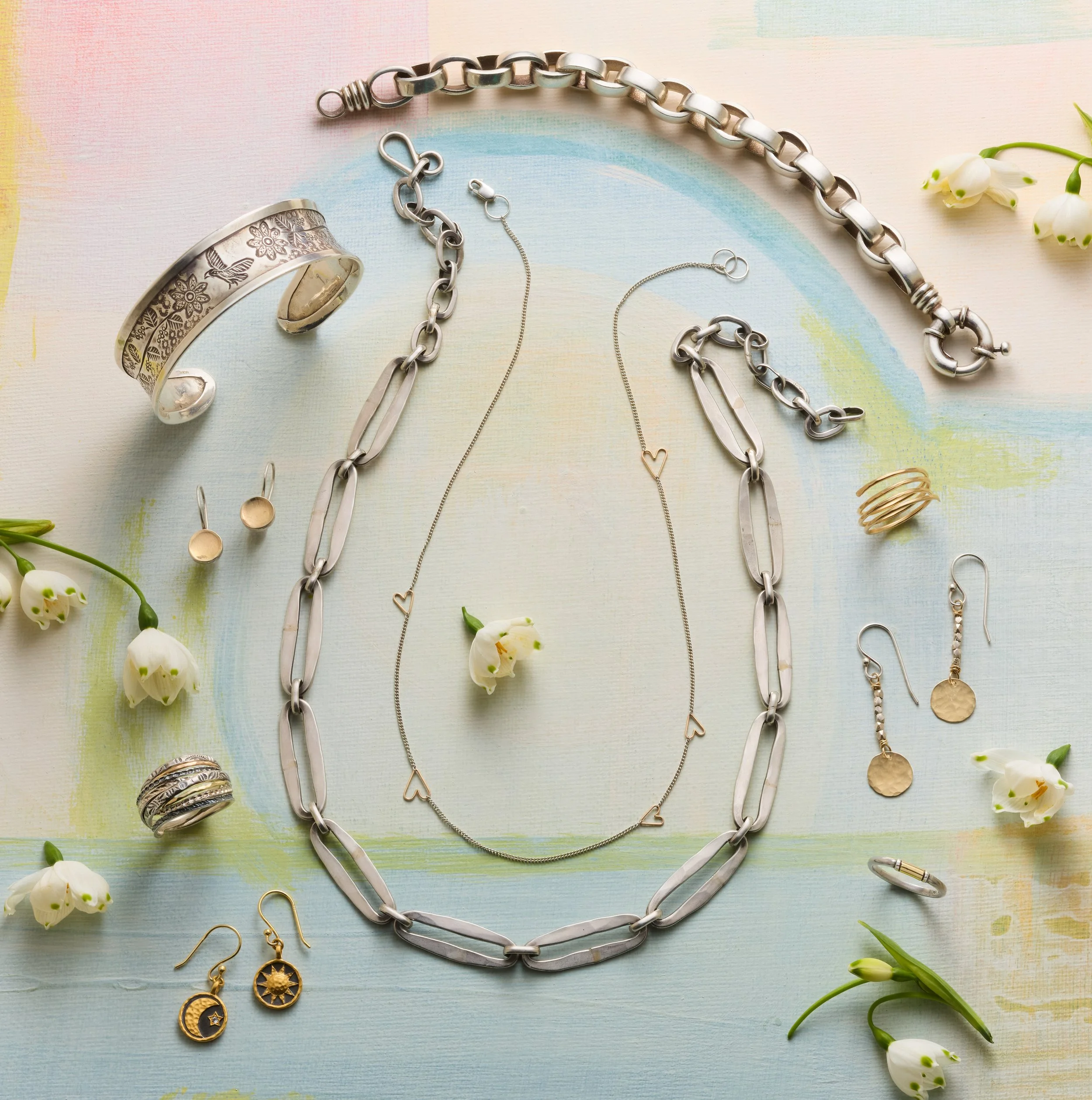Silver and gold jewelry including necklaces, bracelets, earrings, rings, and pendants arranged on a pastel-colored background with white flowers.