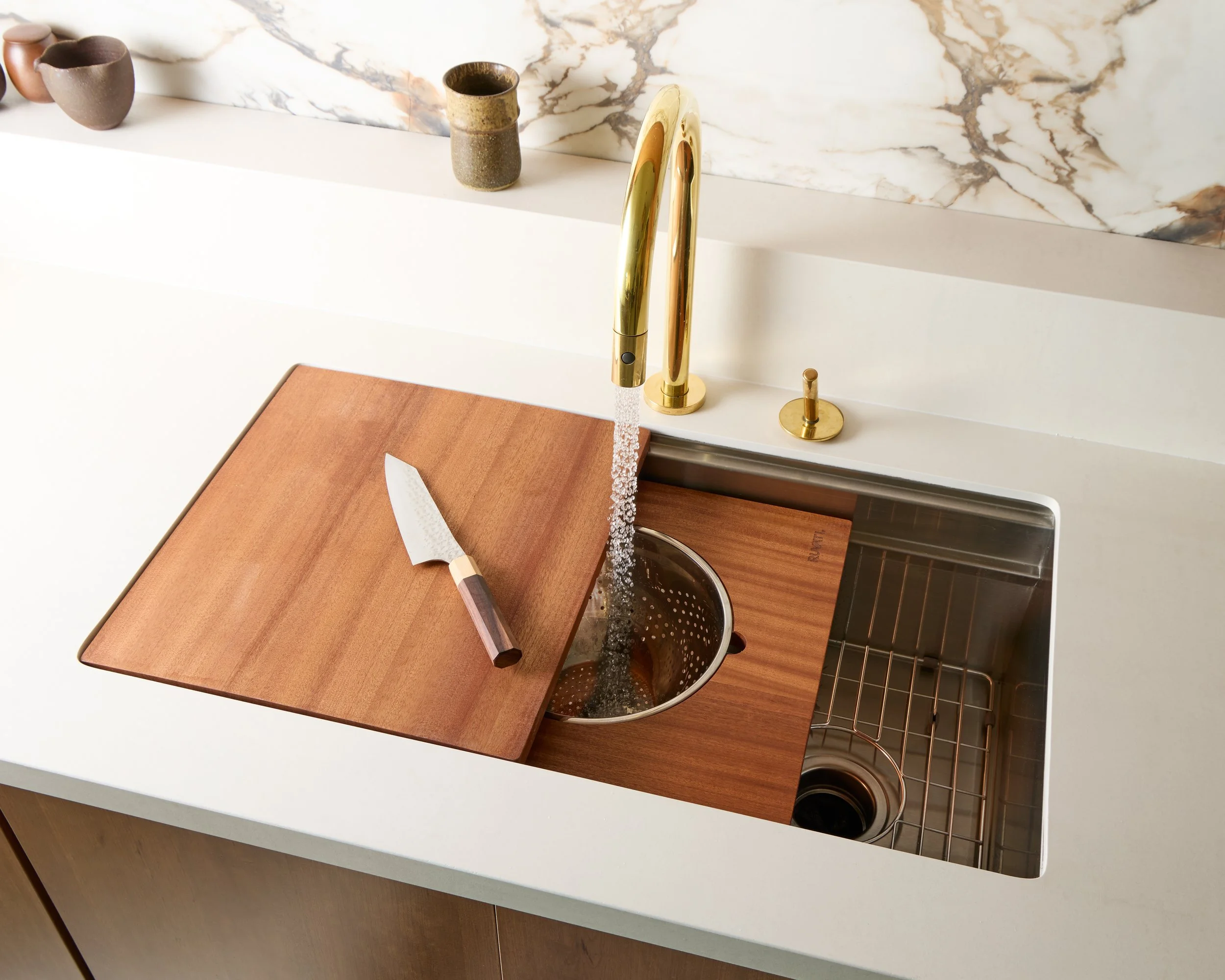 Kitchen sink with a wooden cover, a cutting board and a knife, gold faucet with running water, and marble backsplash.