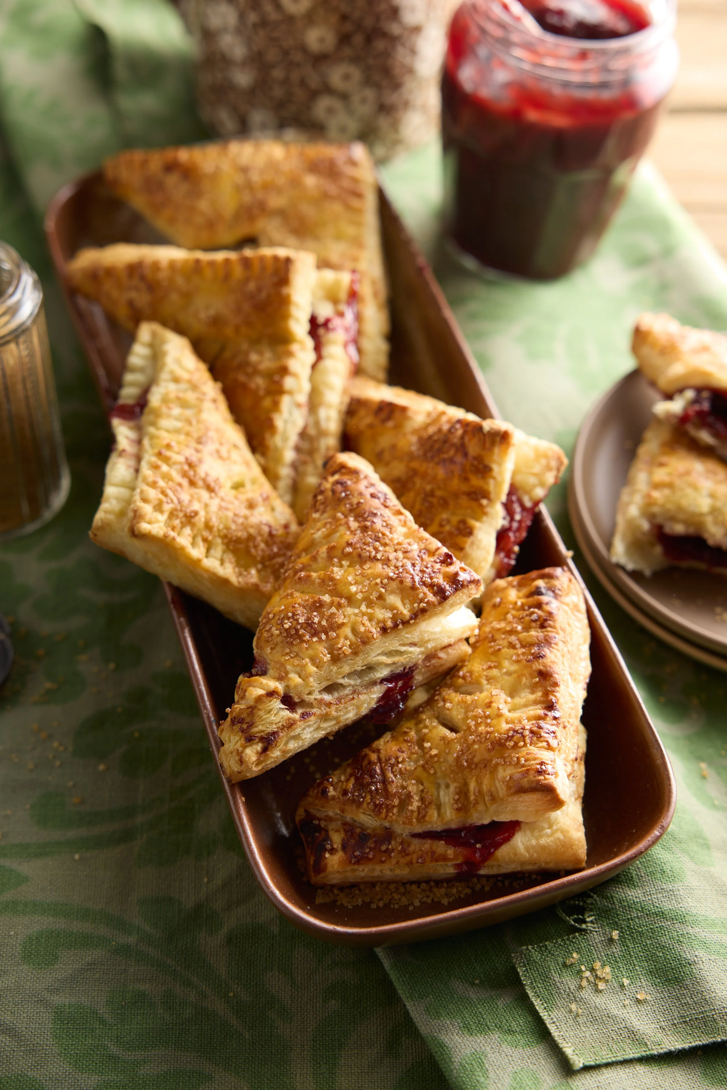 A rectangular dish filled with triangular slices of cherry pie, with a golden, crumbly crust, on a green tablecloth with a jar of cherry jam and a slice of cherry pie on a plate in the background.