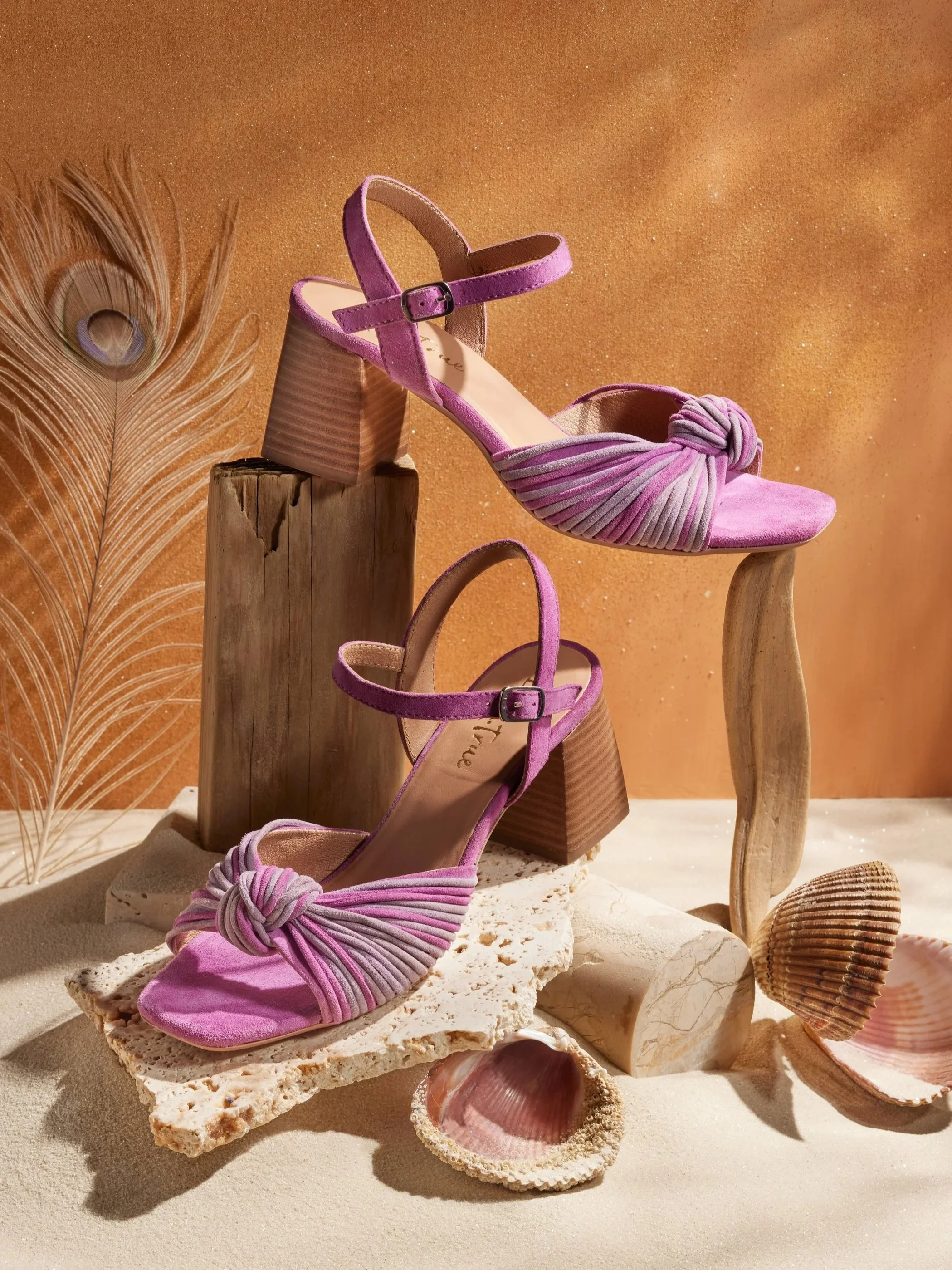 Pink high-heeled sandals with knotted fabric straps, displayed on various wooden and stone props, surrounded by seashells and a peacock feather, against a sandy and warm-toned background.