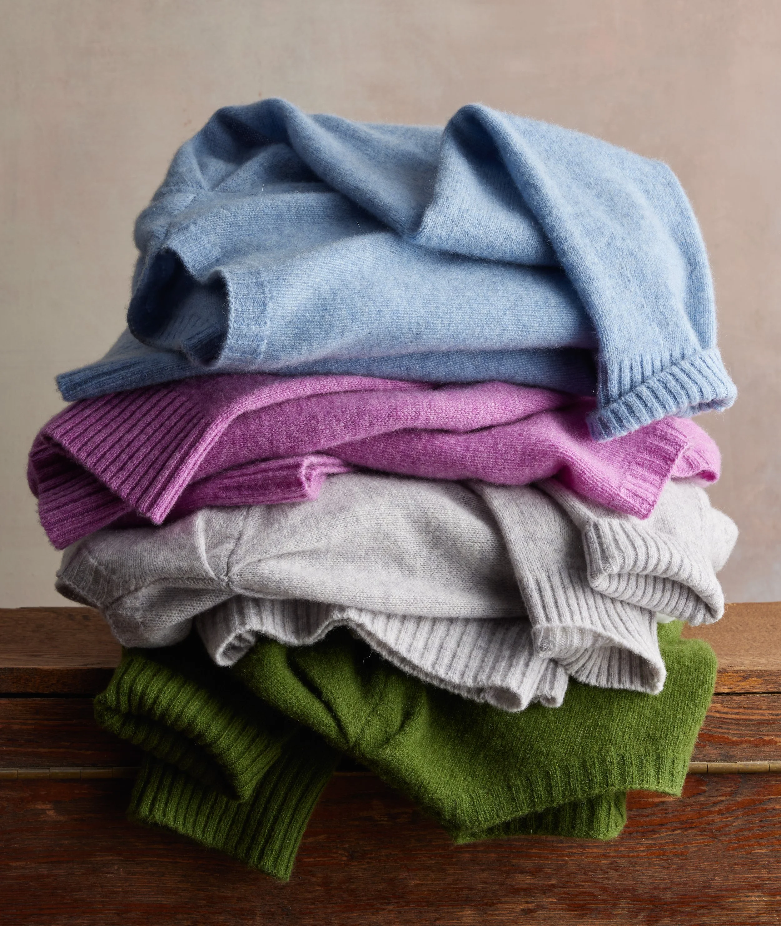Piled stack of folded sweaters in blue, pink, beige, and green colors on wooden surface.