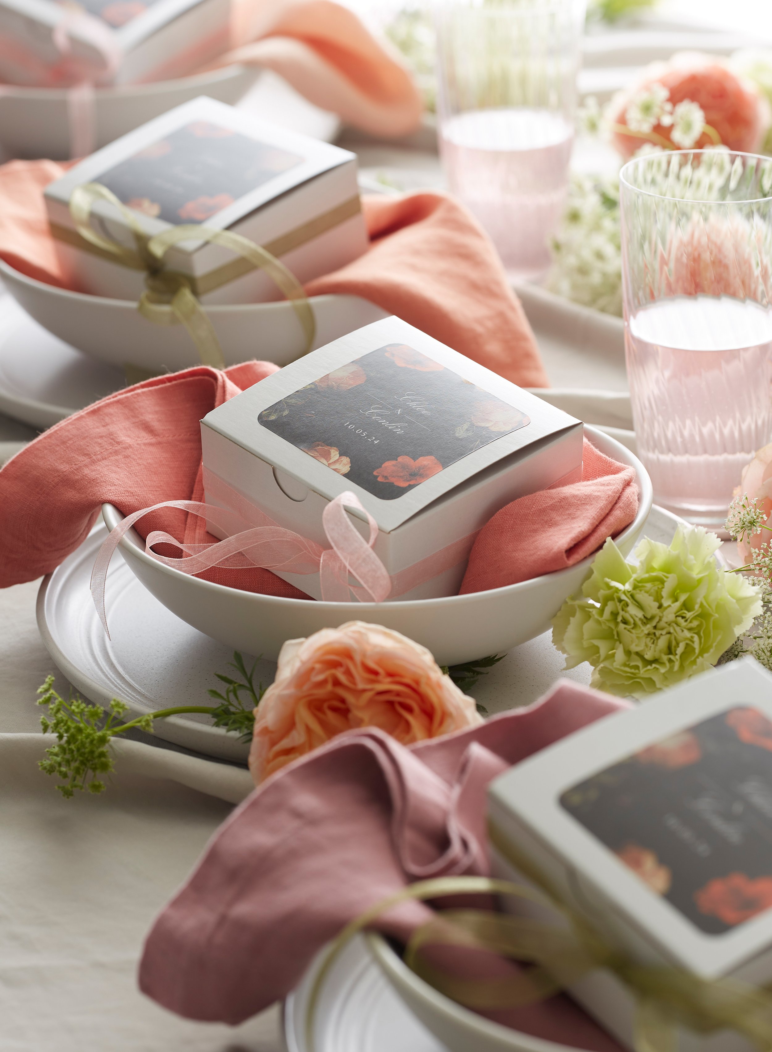 A table setting with pink and peach flowers, napkins, and favor boxes with floral labels, decorative ribbons, and glasses of pink drink.
