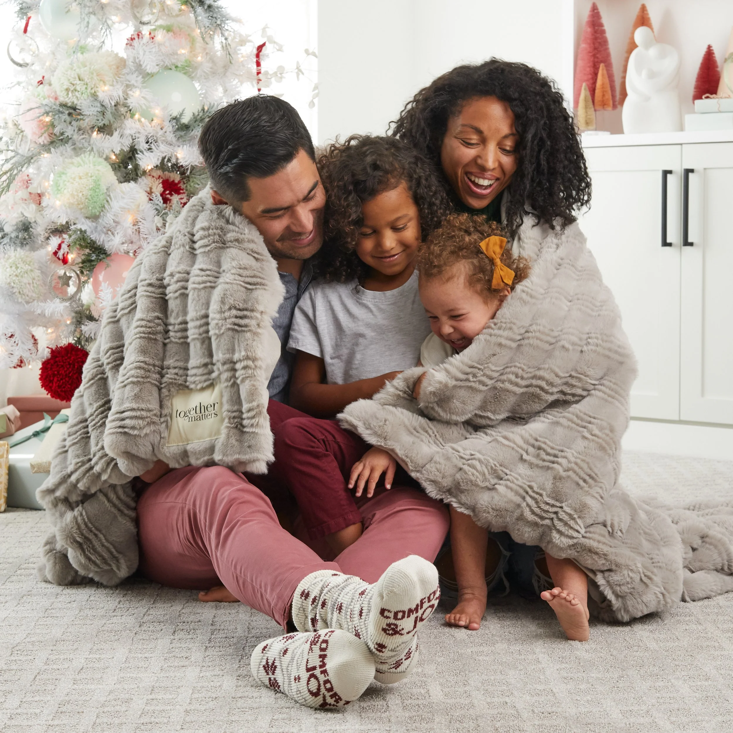 A family of five sitting on a carpeted floor near a decorated Christmas tree, laughing and hugging each other wrapped in cozy blankets.
