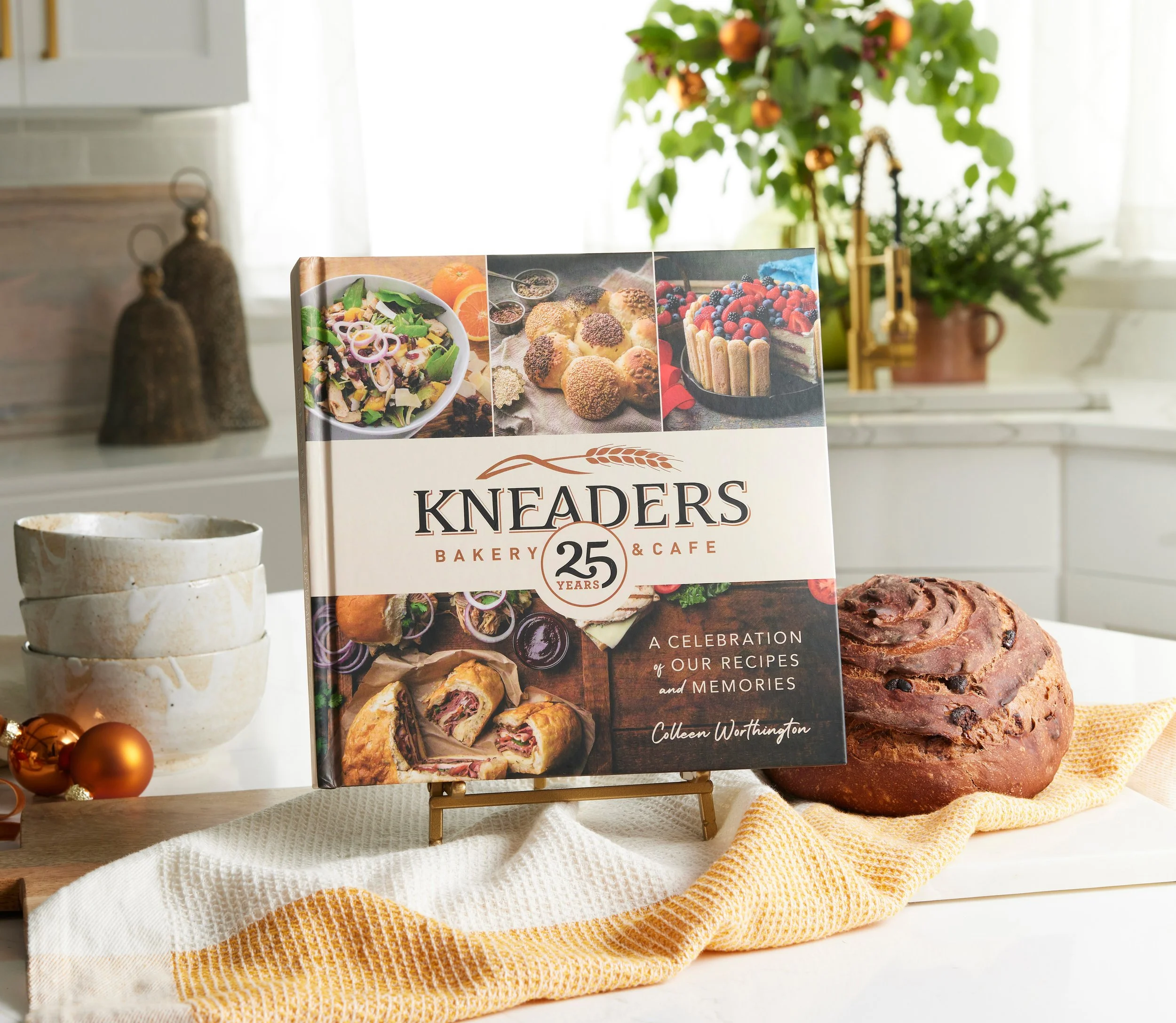 A cookbook titled 'Kneaders Bakery & Cafe, 25 Years' standing on a small easel, with a loaf of chocolate chip bread and a white and orange striped cloth in front; bowls on the left; fairy light ornaments on the left side of the image, and a potted plant and kitchen window in the background.