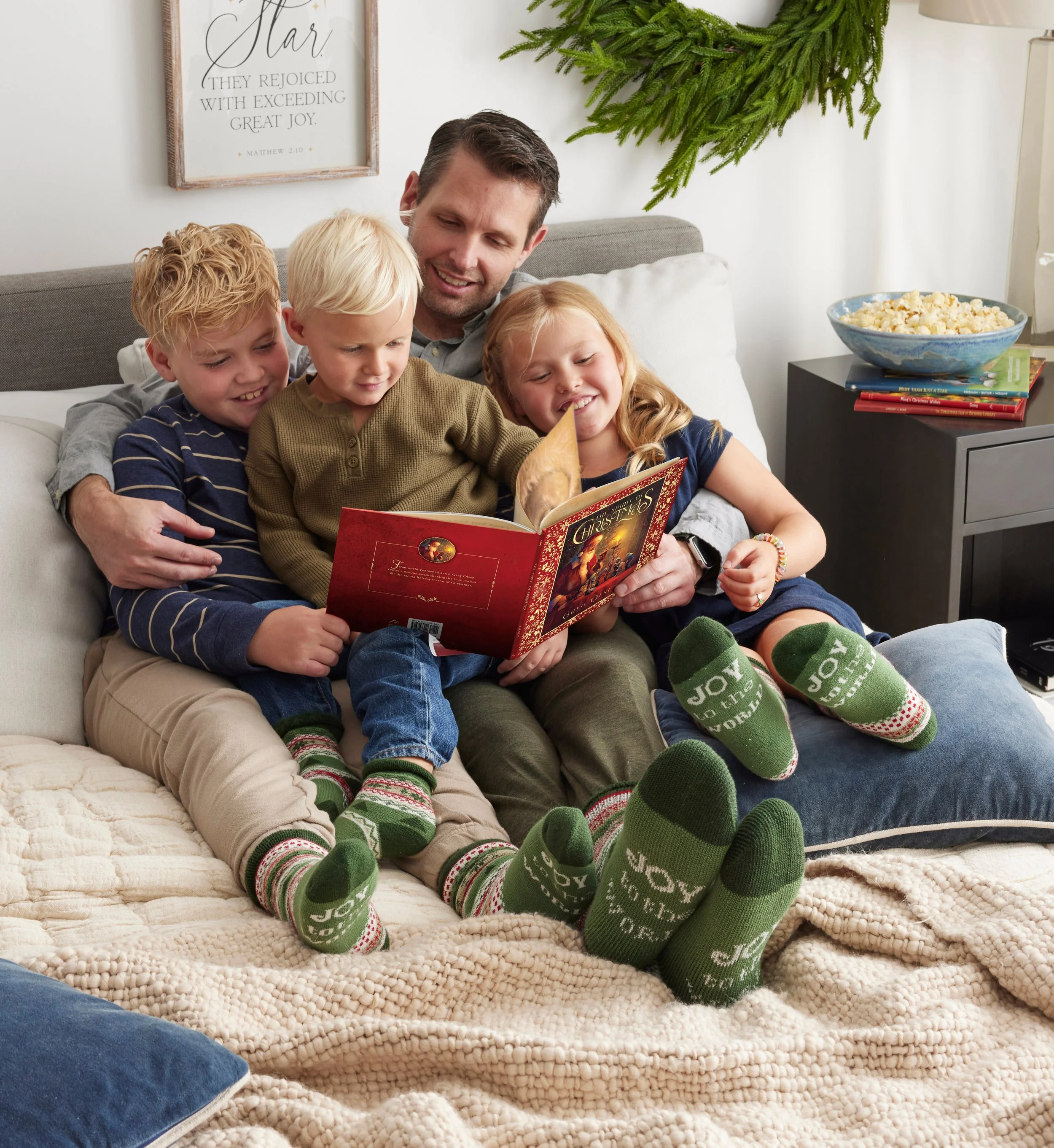 A family of five, including a man and four children, sitting on a bed and reading a Christmas book together while wearing green socks with holiday messages. They are smiling and enjoying a cozy moment.