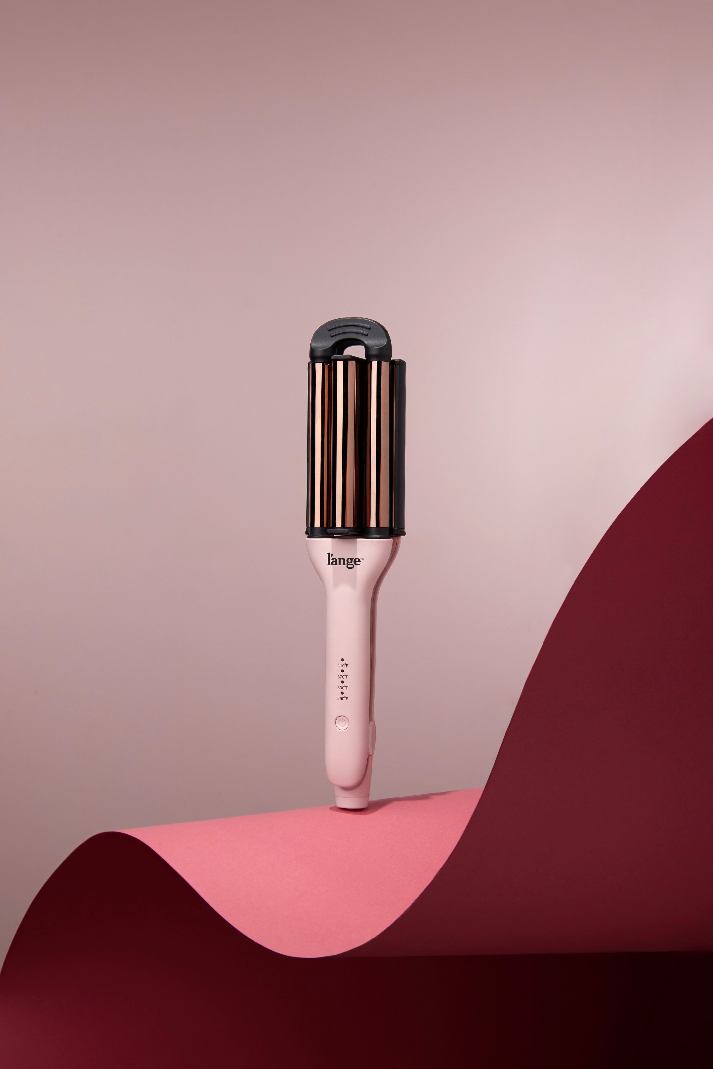 A pink and black hair straightener or curling iron standing upright on a pink surface, with a gradient background from light pink to beige.