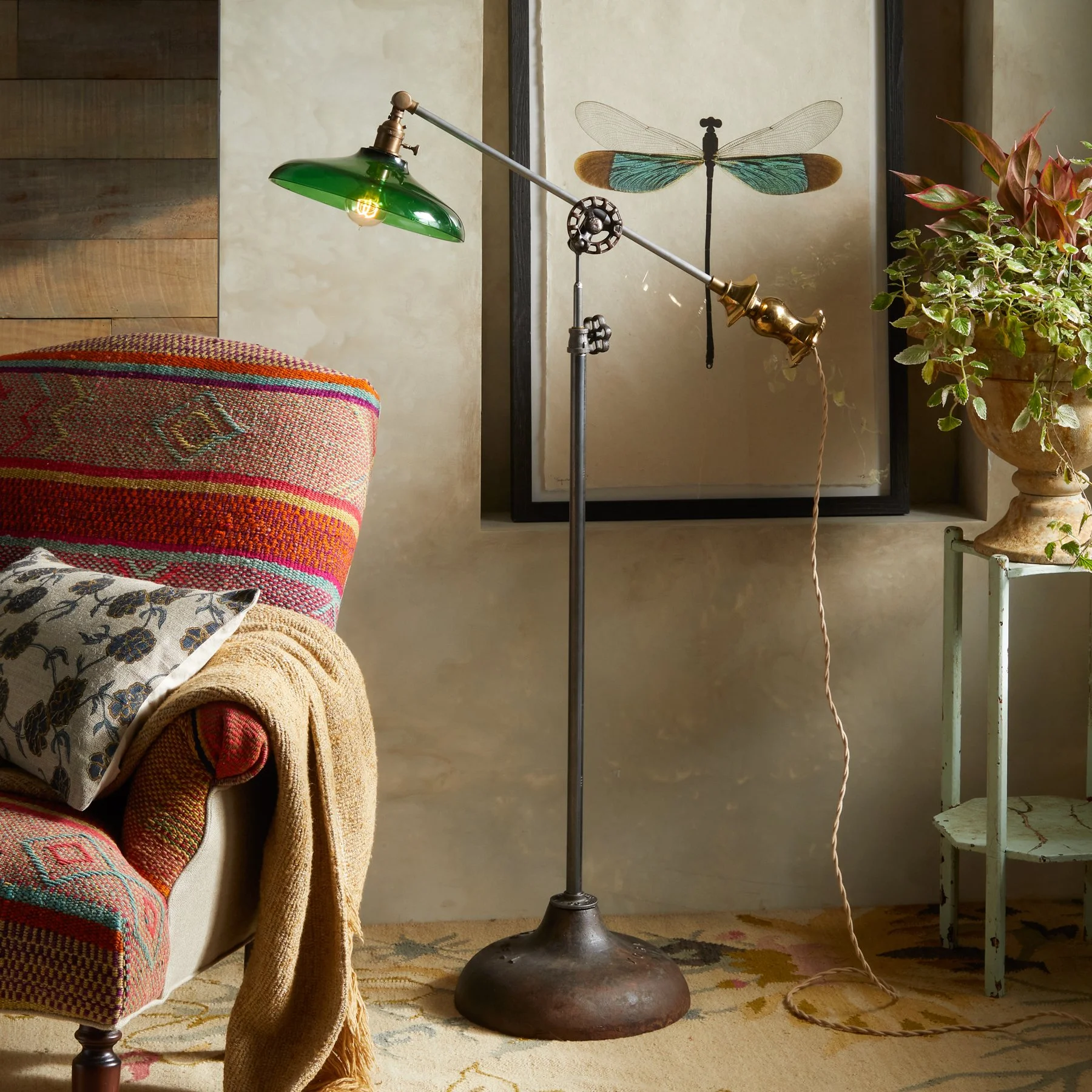 A vintage-style floor lamp with a green shade, brass accents, and an adjustable arm, standing next to a sofa and a framed dragonfly artwork in a cozy living room.