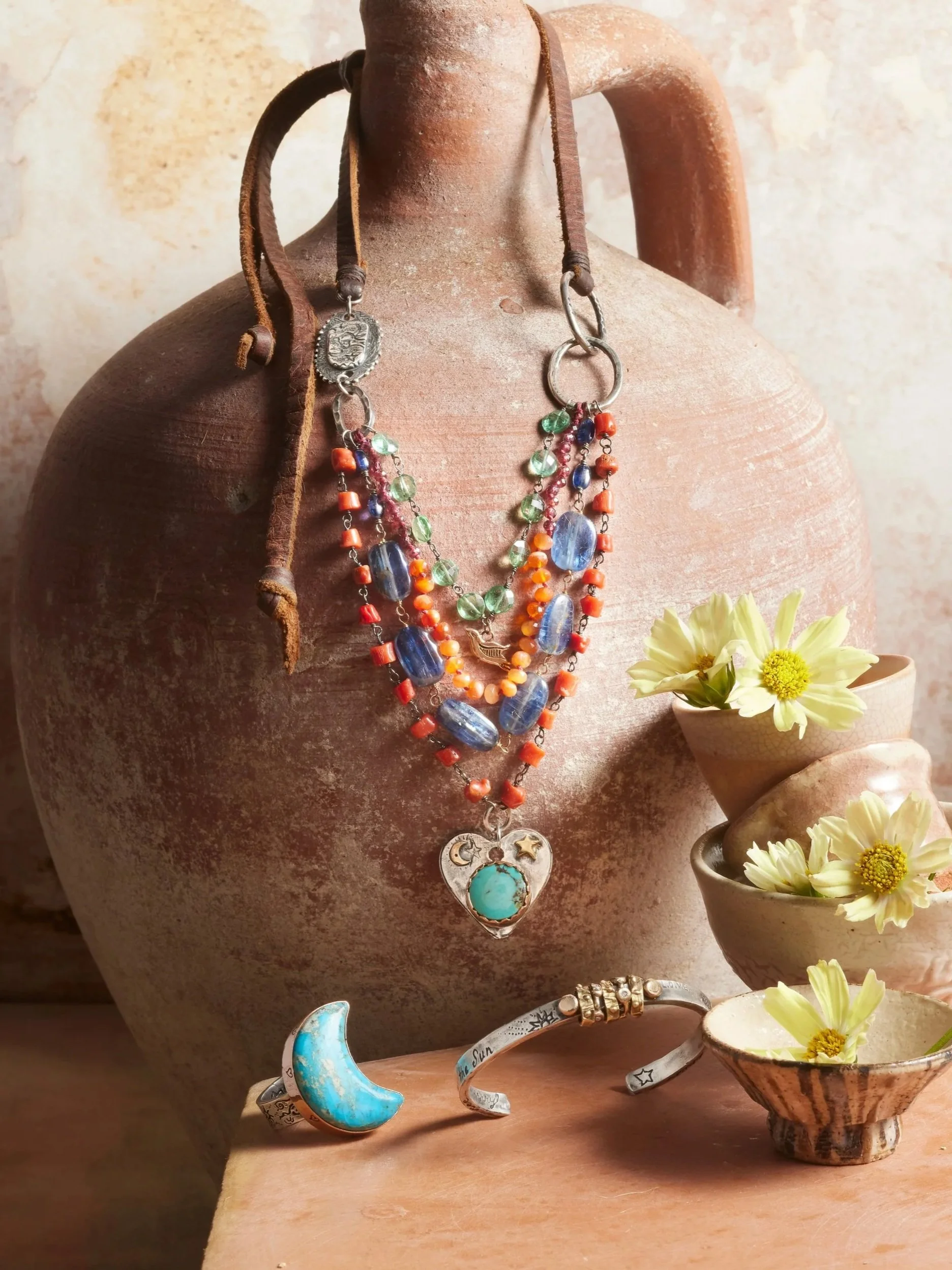 Display of colorful jewelry including layered necklaces with beads and charms, a moon-shaped bracelet, and a turquoise heart pendant, arranged on a rustic ceramic vessel with small cups of yellow flowers nearby.