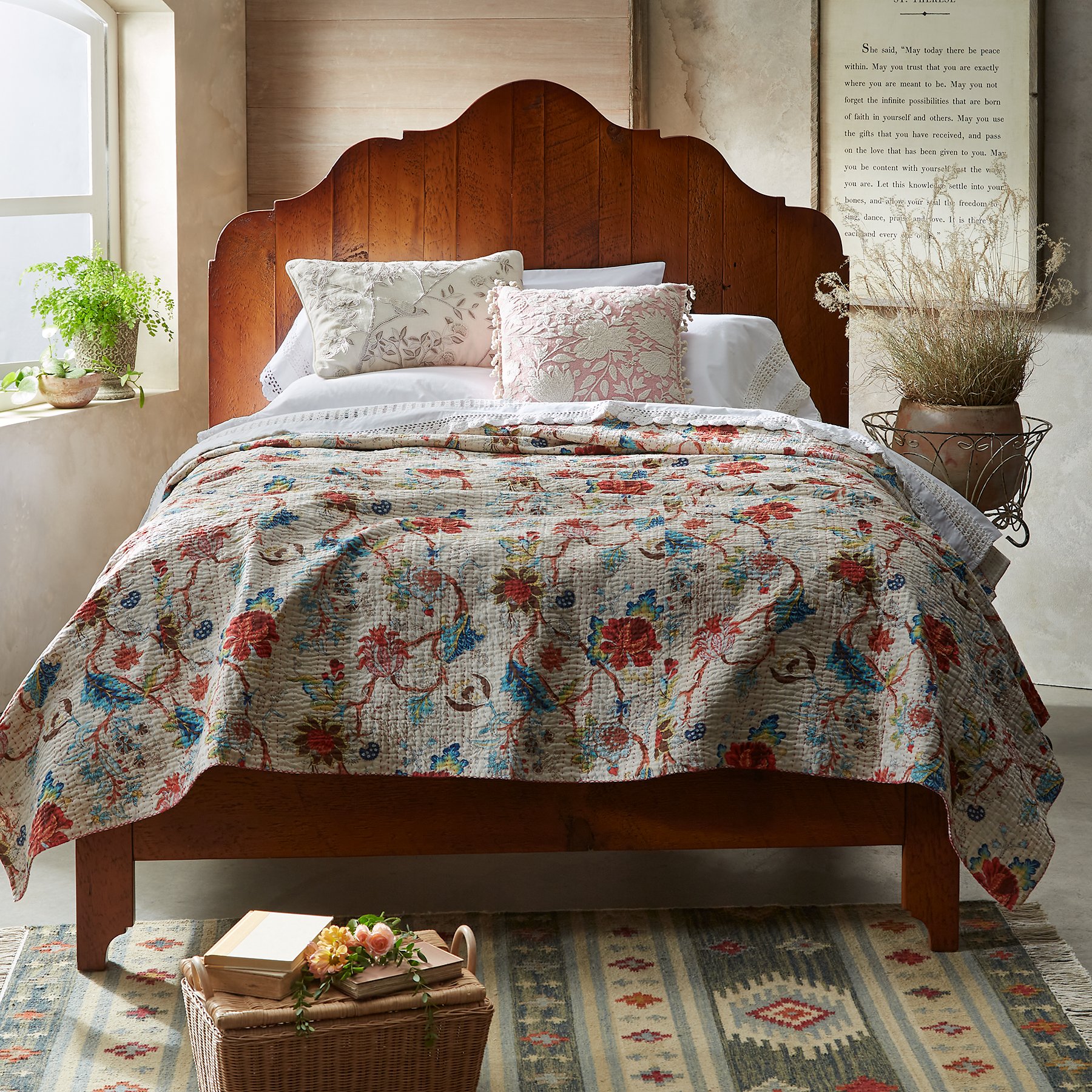 A cozy bedroom with a wooden bed frame, floral quilt, decorative pillows, plants on the window sill and beside the bed, and a woven basket with books and flowers on a patterned rug.