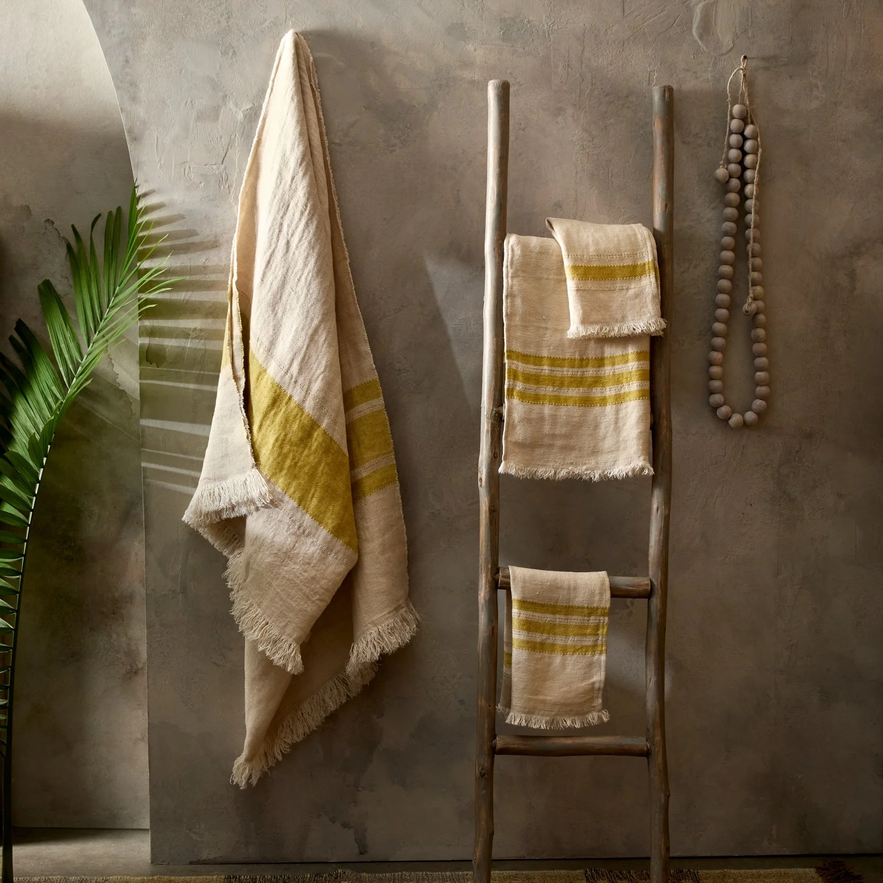 Towels hanging on a wooden ladder and a wall, with a green plant shadowed on the left side and a beaded ornament hanging on the wall, against a textured wall background.
