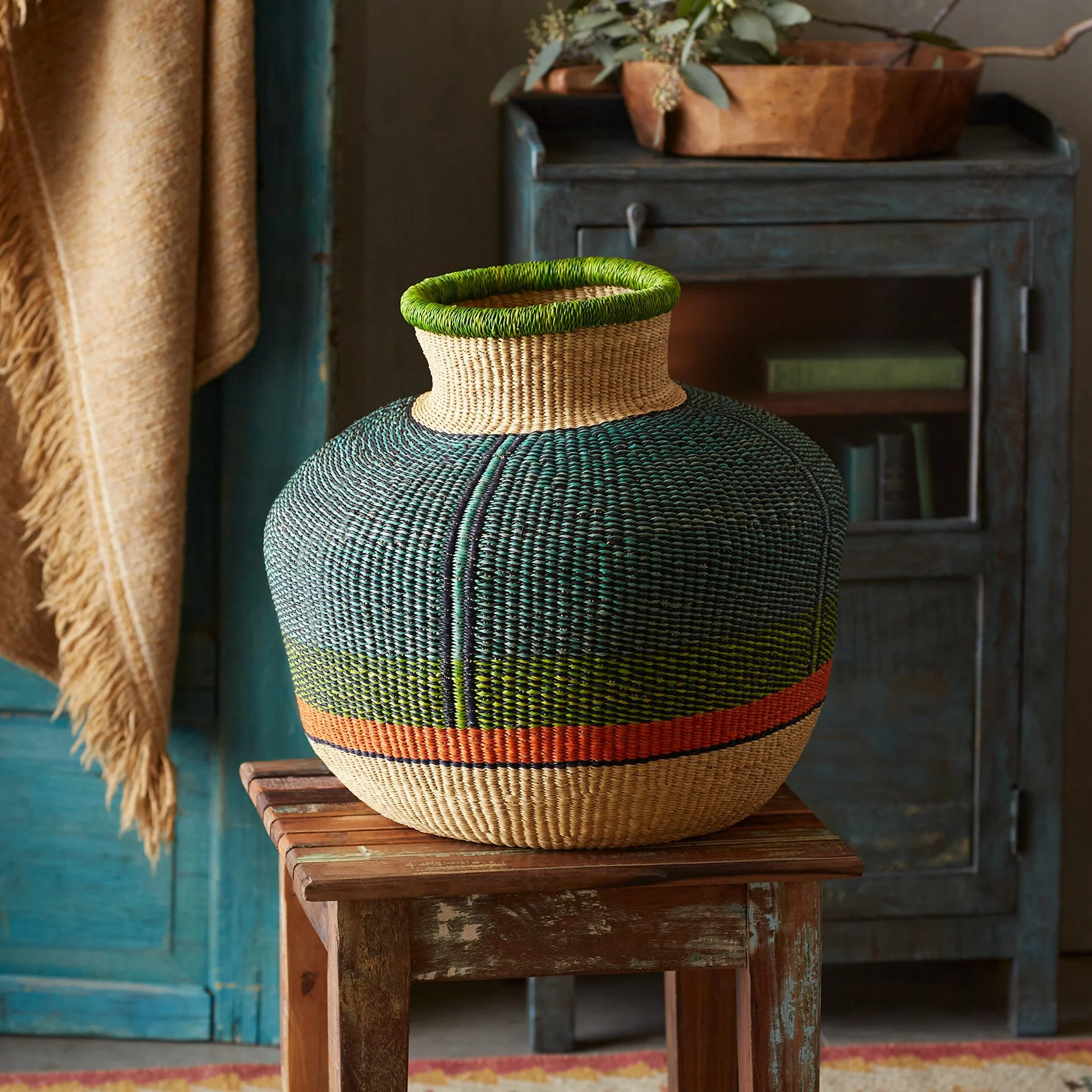 A colorful woven basket with a round shape, featuring a pattern of green, black, beige, and orange colors, sitting on a small wooden stool in a rustic room.