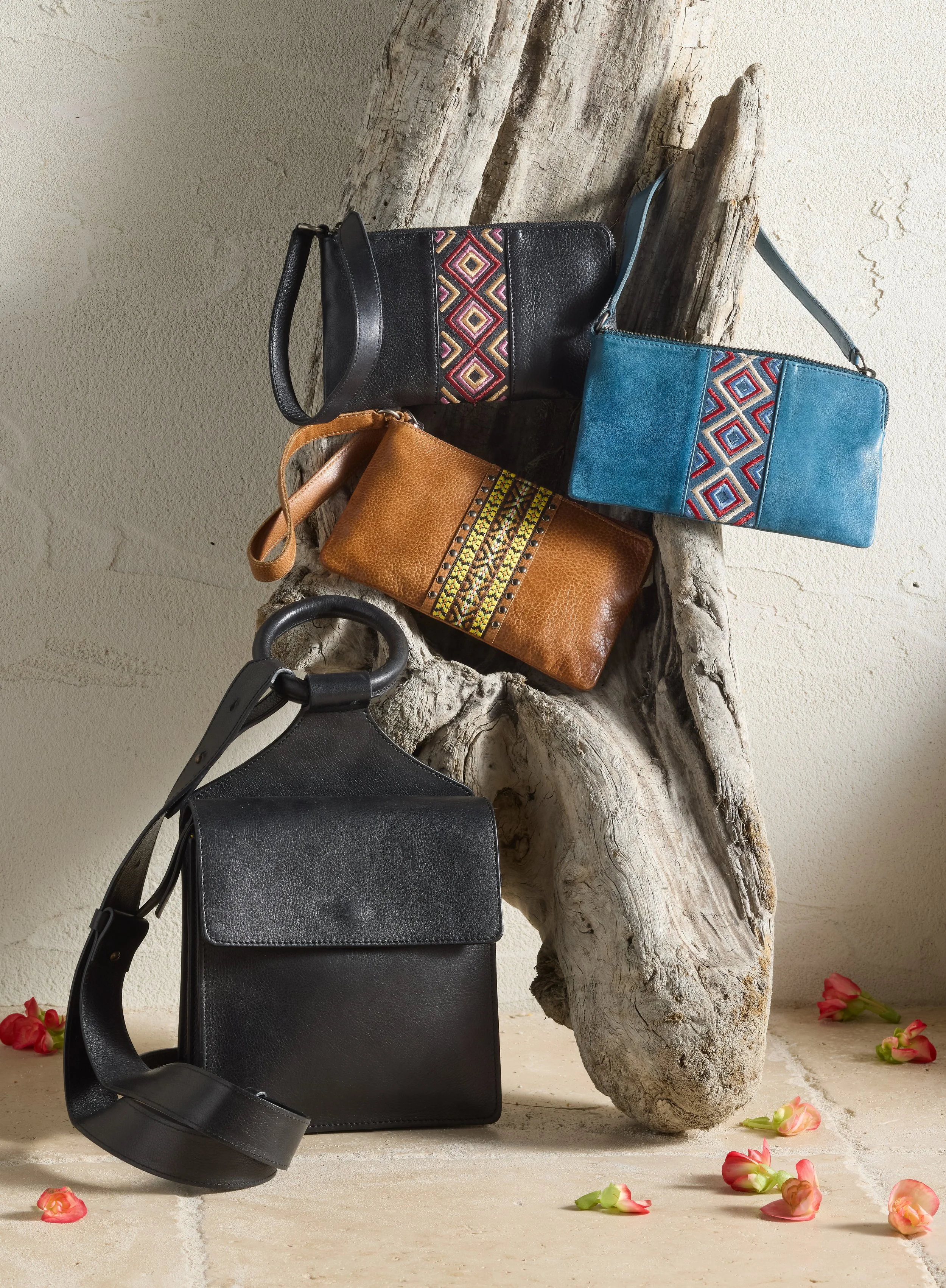 Four handbags, three with colorful geometric patterns and one plain black, arranged on a piece of driftwood with small pink flowers scattered around the floor.