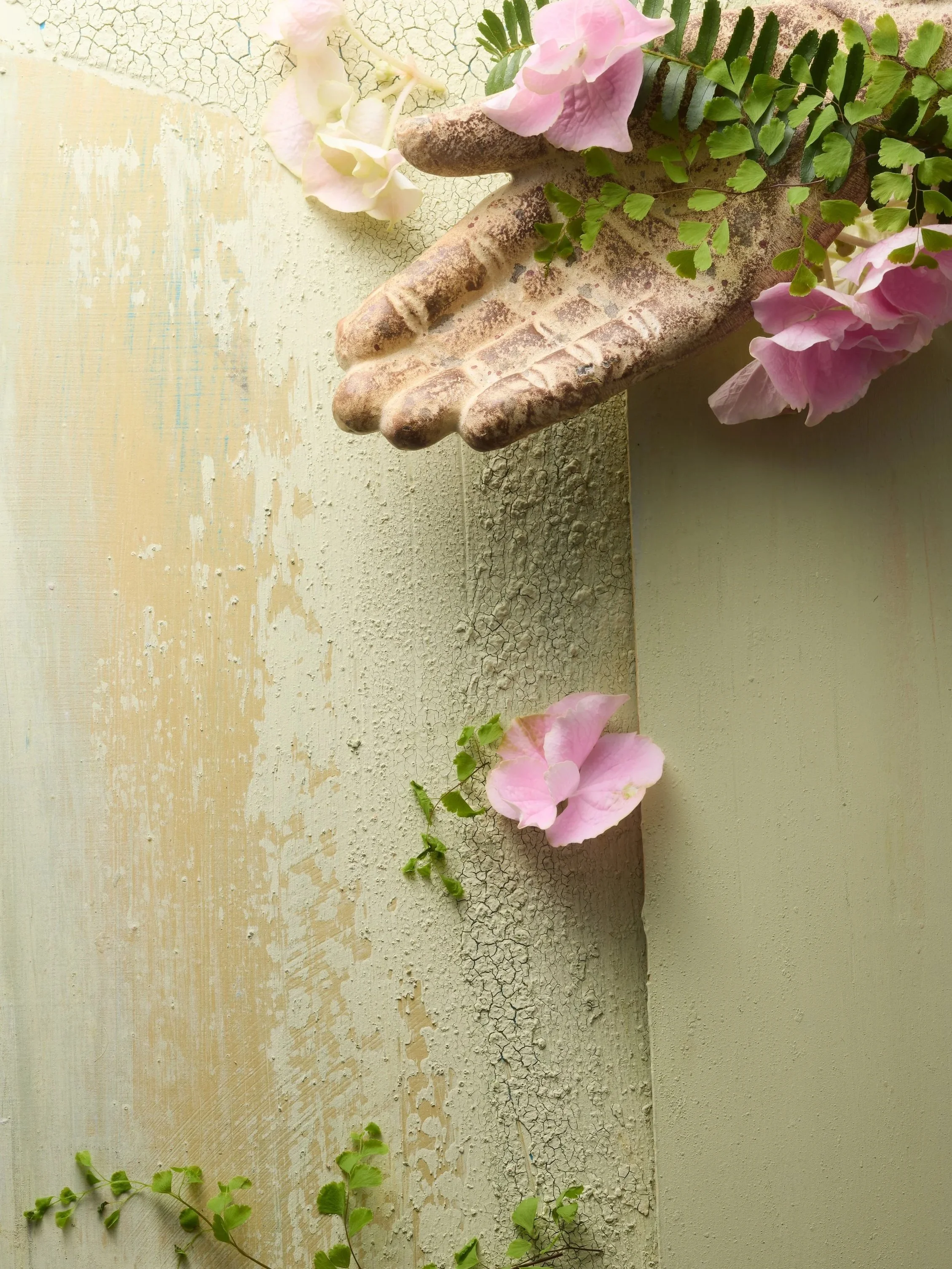 A weathered, decorative stone hand with fingers extended, holding pink flowers and green foliage against a textured beige wall.
