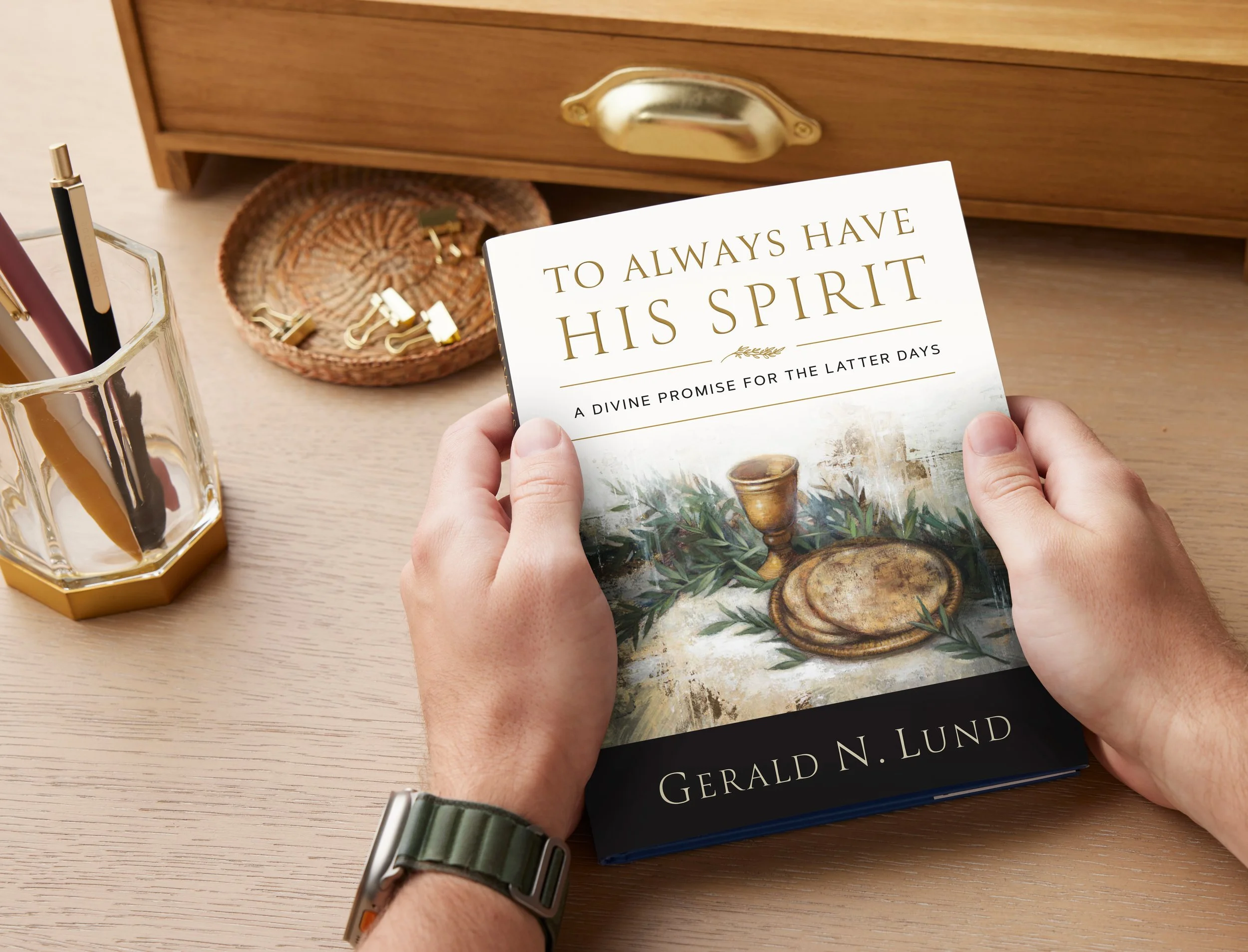 A person holding a book titled 'To Always Have His Spirit' by Gerald N. Lund, on a wooden desk with a glass container of pens and markers, a wicker basket with gold paper clips, and a wooden drawer in the background.