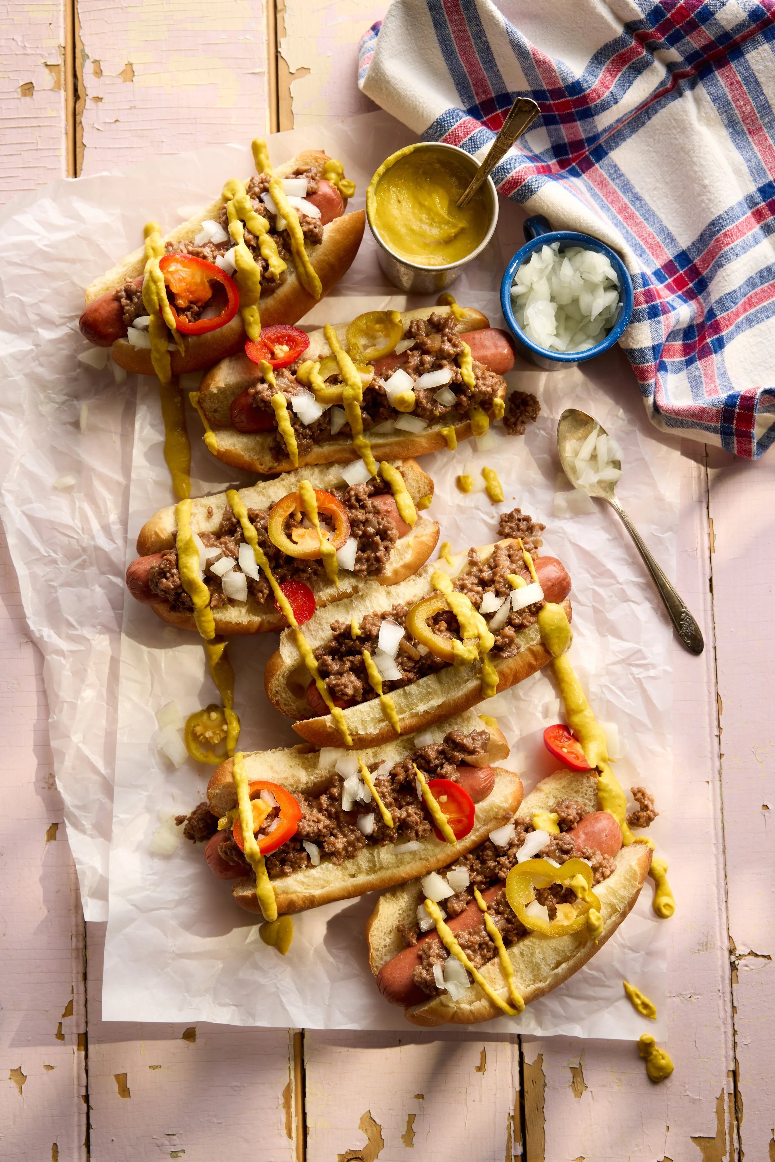 Hot dogs topped with mustard, onions, and chili, served on a sheet of parchment paper with condiments on the side.