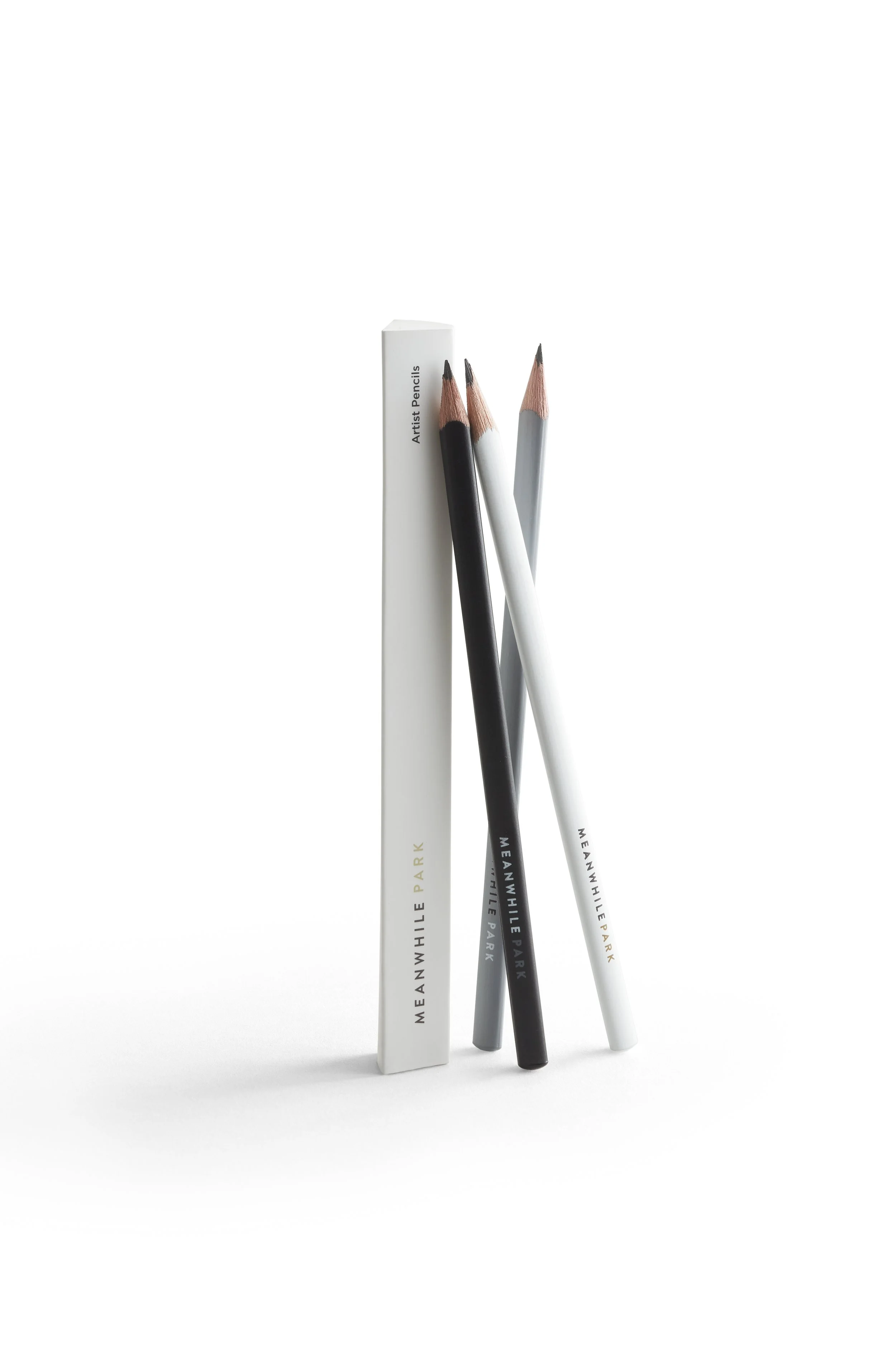 Three artist pencils in black, white, and gray, leaning against a white box labeled 'Artist Pencils' with 'MEANWHILE PARK' printed on the pencils, on a white background.