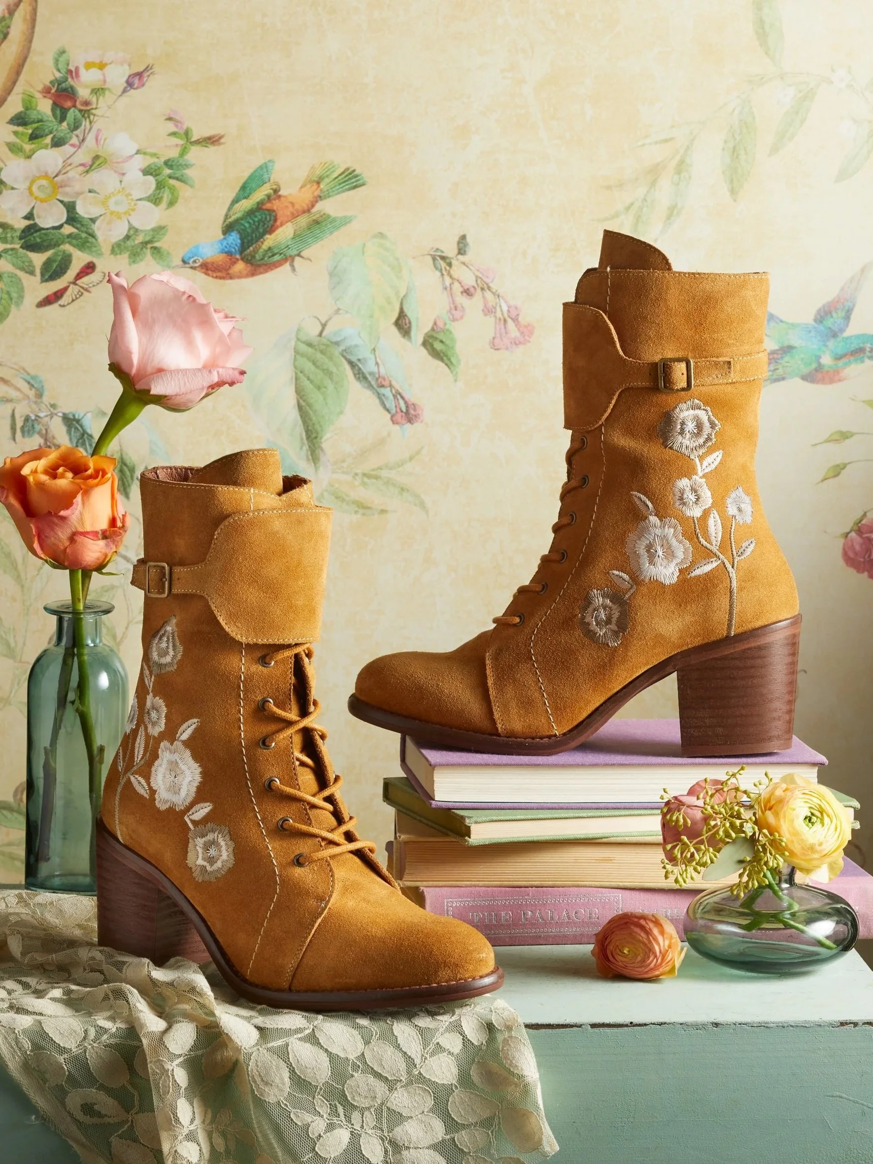 A pair of tan suede lace-up ankle boots with embroidered white flowers, placed on pastel-colored books, with flowers in glass vases and a floral wallpaper background.