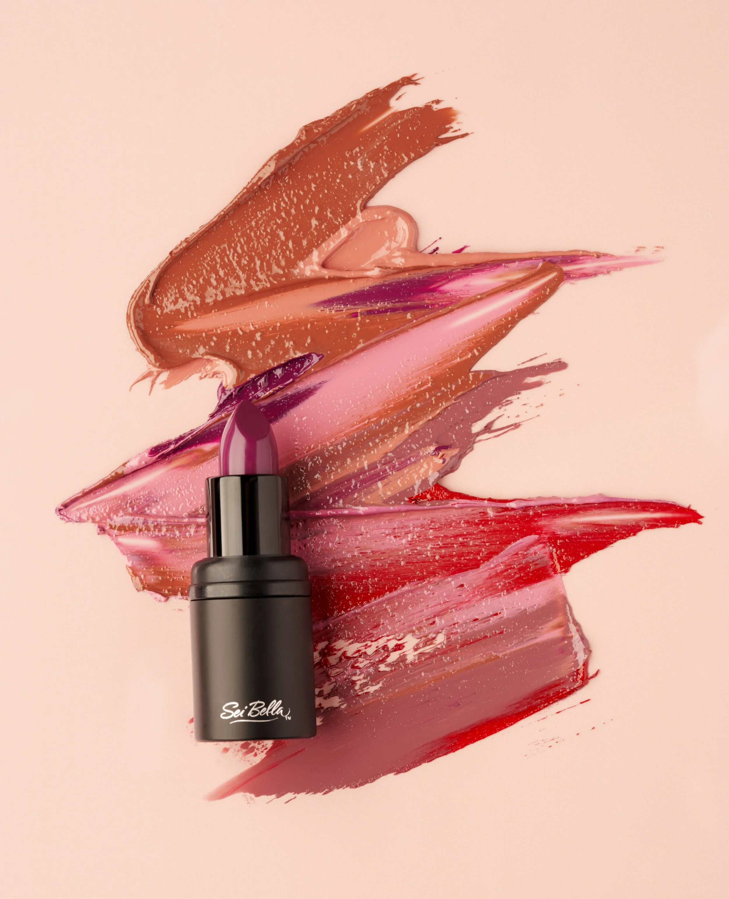 Makeup brush with pink lipstick smear and various shades of lipstick in red, pink, beige, and purple on a light pink background.