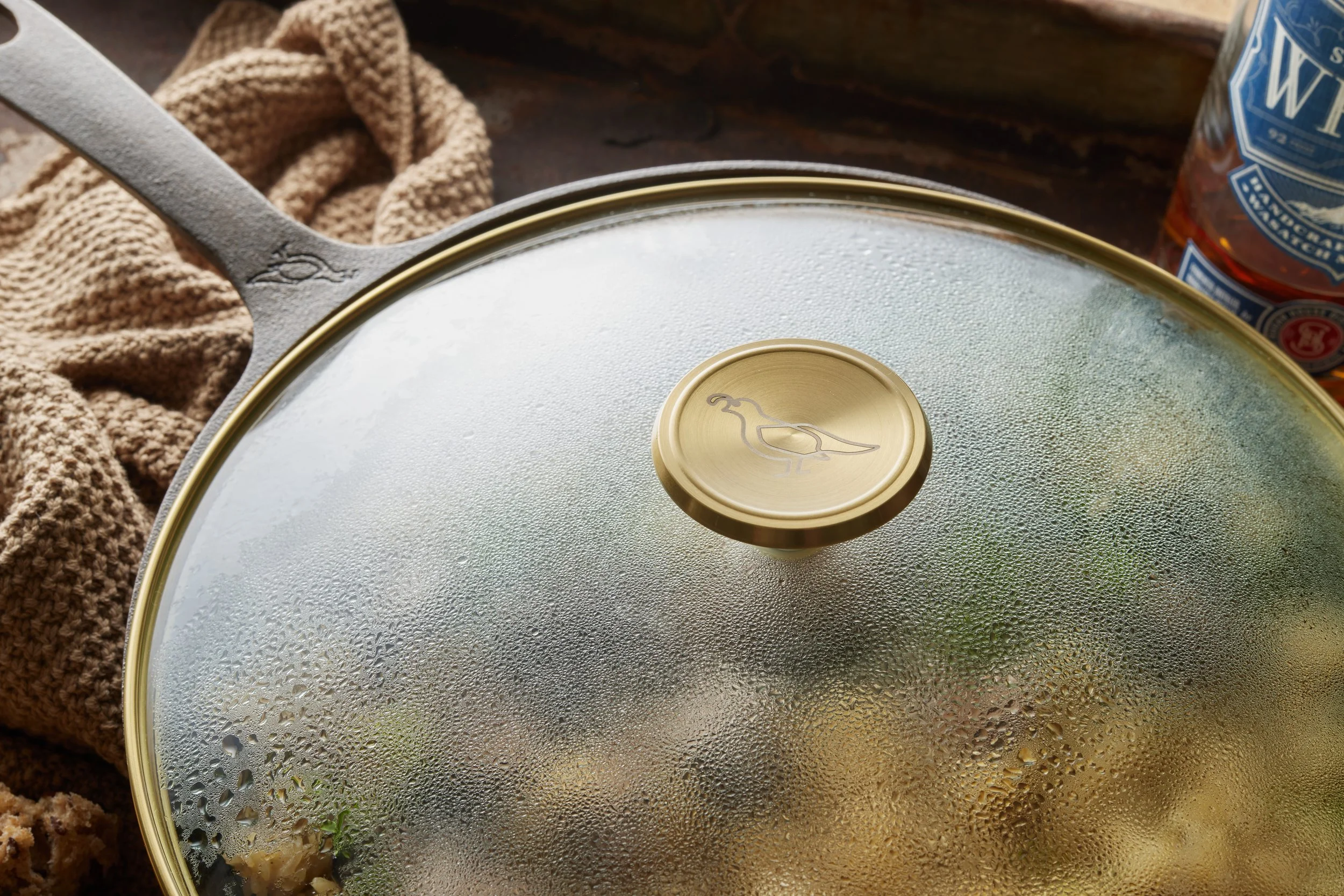 Glass lid with a gold rim and a bird logo in the center, condensation on the outside, placed on a pan with a wooden surface and a beige cloth nearby, and a bottle with blue and white label in the background.