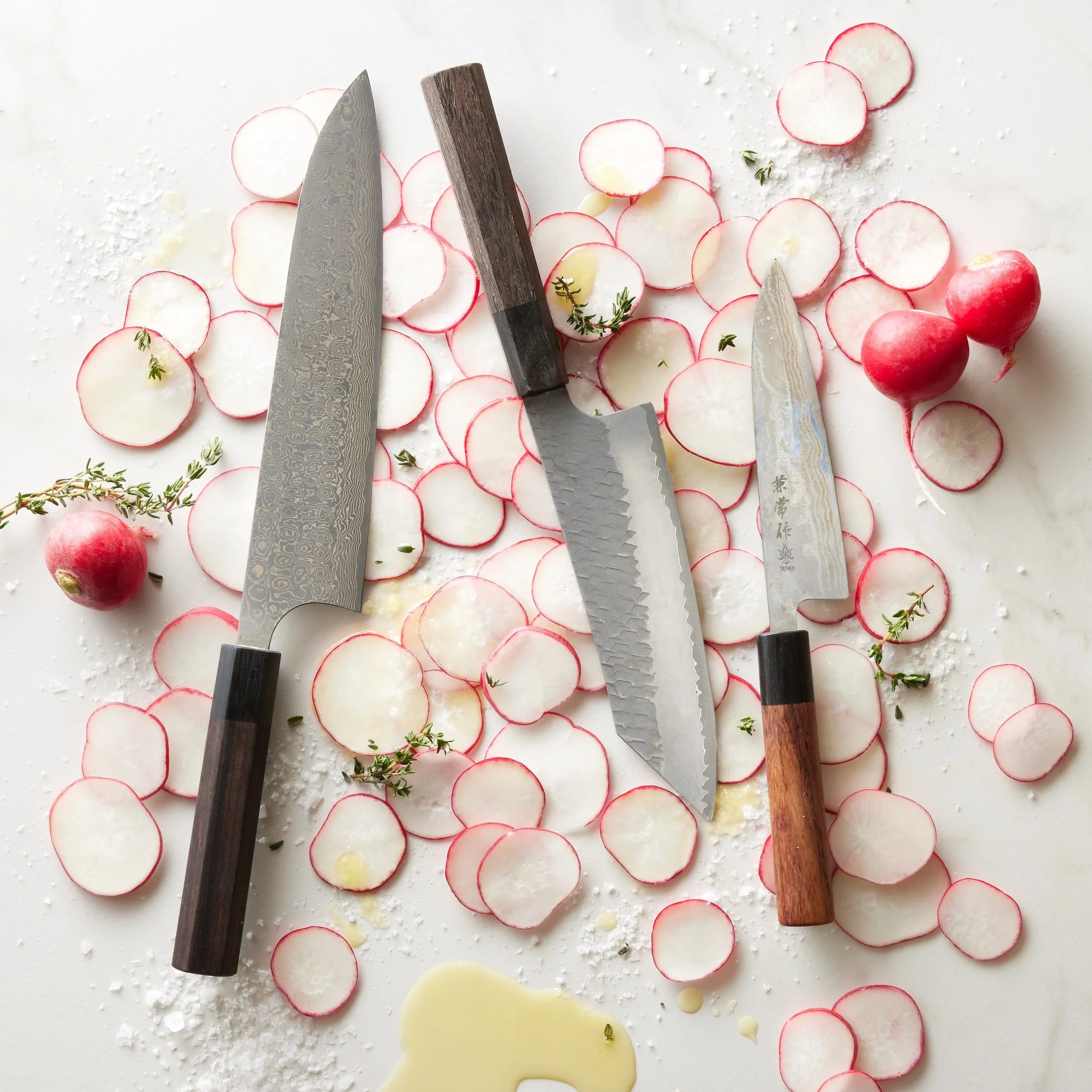 Sliced radishes on a white surface with three knives, fresh thyme sprigs, and whole radishes.