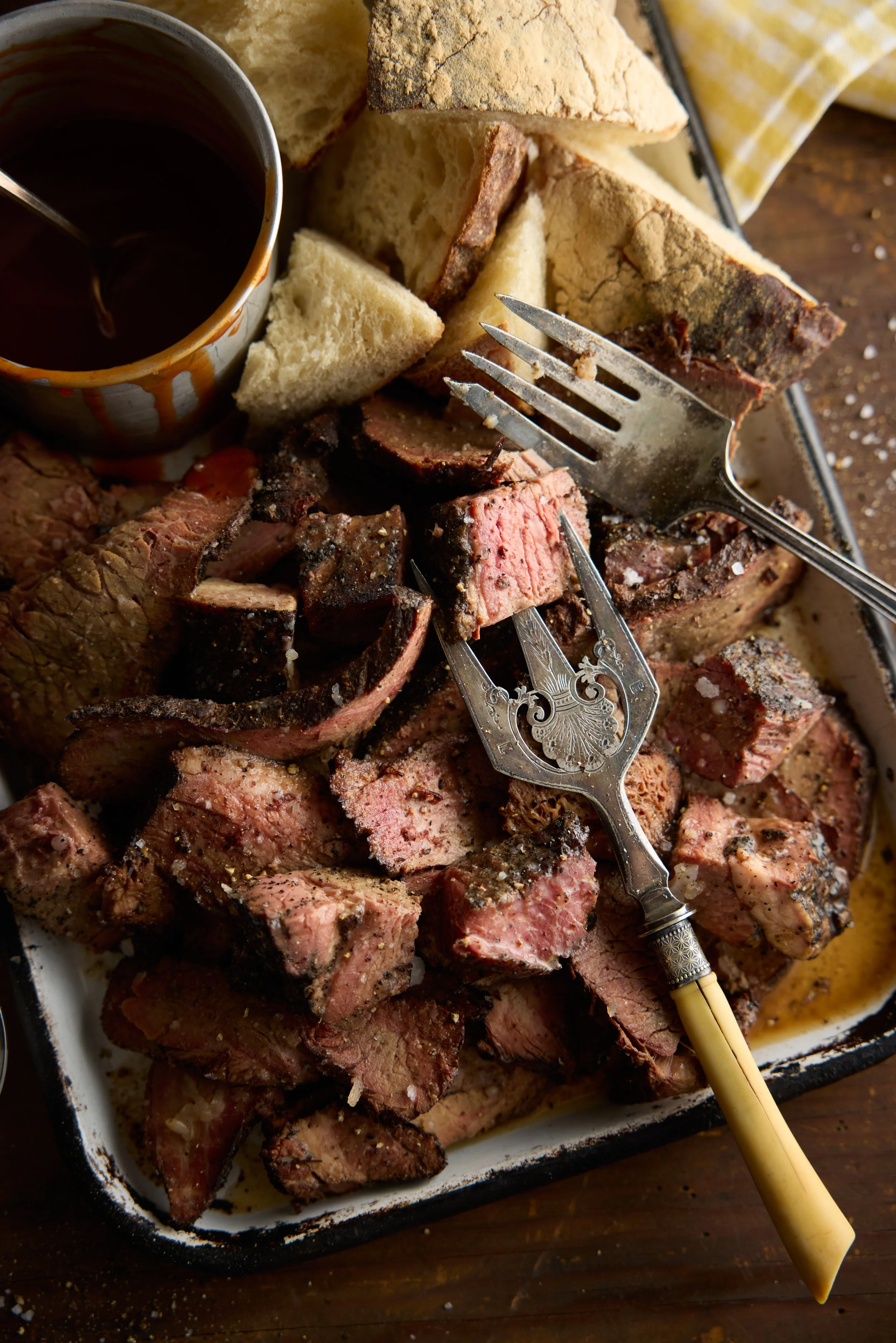 A tray of assorted cooked steaks with a cup of dark gravy or sauce, pieces of bread, and a yellow checkered cloth in the background.