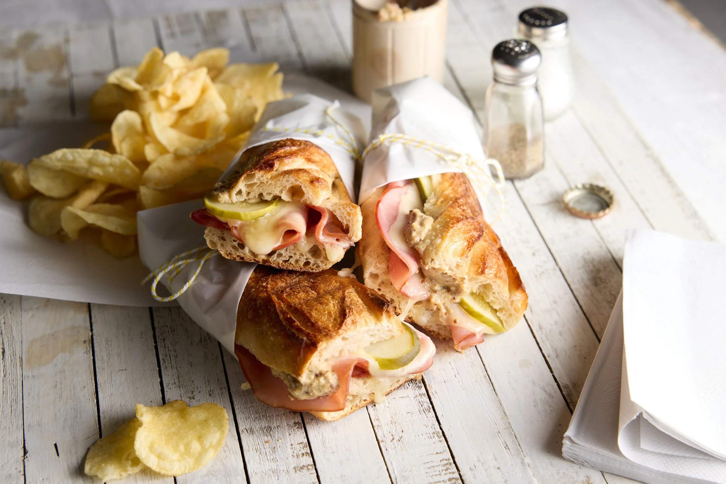 Club sandwich wrap with sliced pickles, ham, turkey, cheese, and lettuce, served with potato chips on a white wooden table.