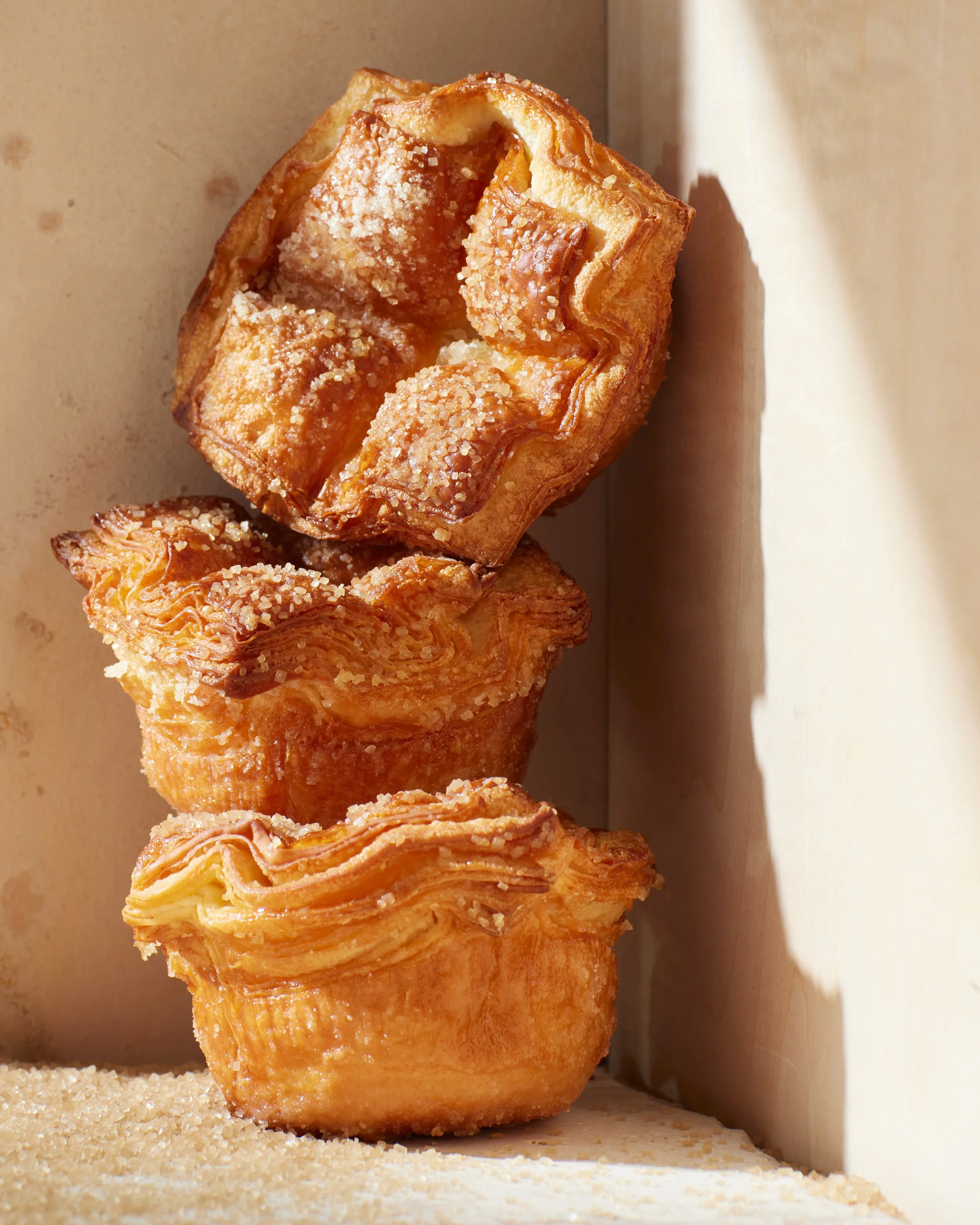 Three golden brown muffins, topped with sugar crystals, stacked on a wooden surface.